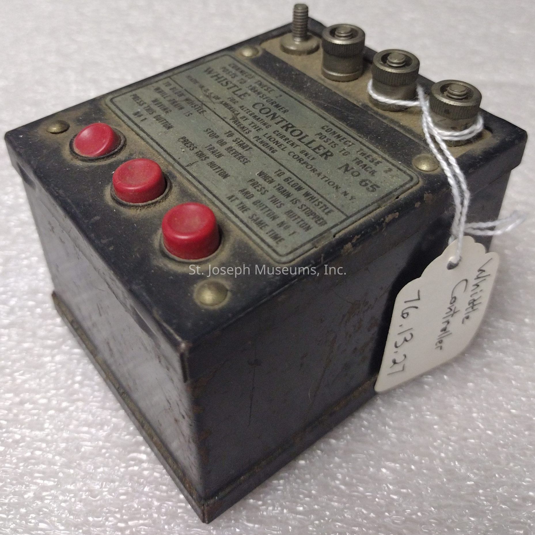 The image shows a vintage whistle controller, labeled as "Whistle Controller No. 65," designed for controlling model trains. It is a rectangular, metal box with a worn appearance, featuring three prominent red buttons on the top surface. The panel displays various instructions and connection details, indicating its use with specific electrical setups. Three metal knobs, tied together with white string, are visible on the top, likely used for wire connections. A paper tag attached to the device reads "Whistle Controller 76.13.27," possibly indicating a catalog or inventory number. The device appears to be resting on a textured, white surface, highlighting its dusty, aged condition.