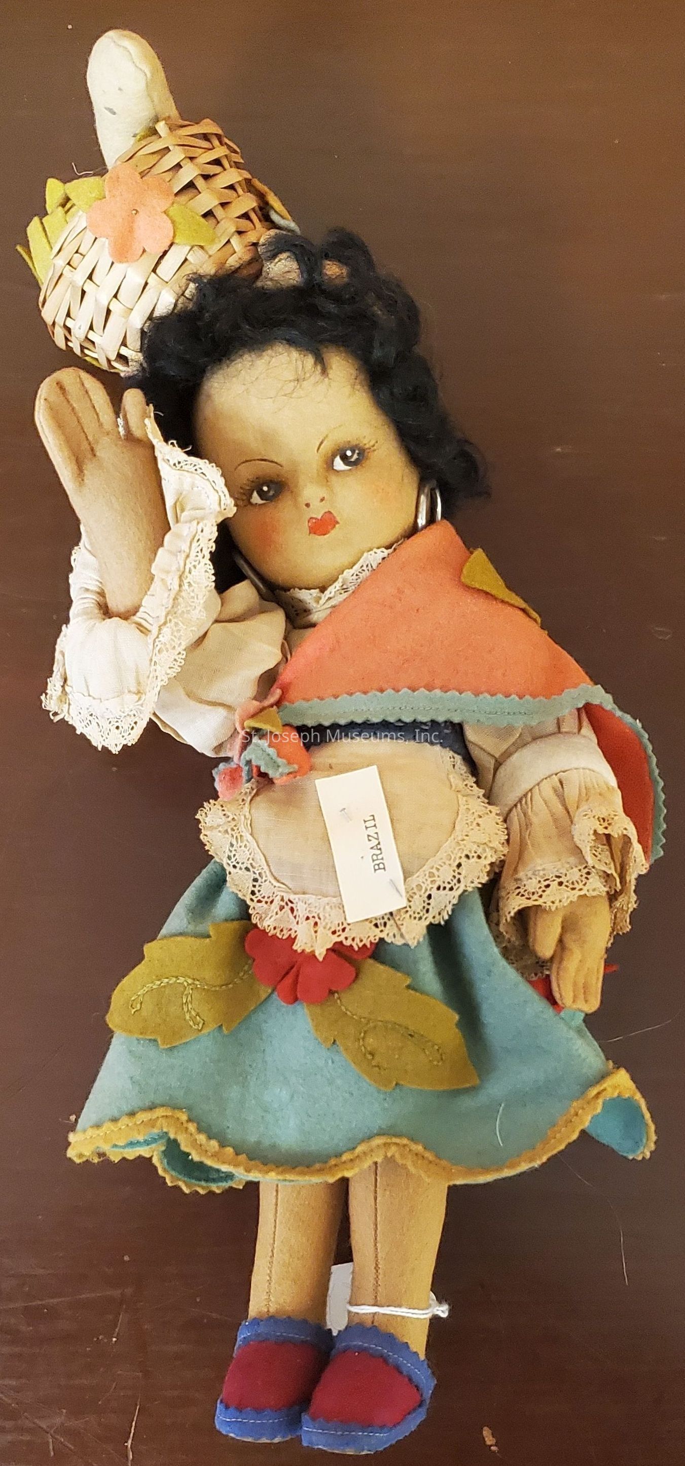 The image displays a charming handcrafted doll with an expressive face, dressed in traditional attire. The doll's outfit consists of a beige blouse adorned with intricate lace, a vibrant orange shawl, and a blue skirt featuring decorative felt leaves and flowers. She wears red and blue shoes, and holds a woven basket on her head, which is highlighted with a fabric flower. The doll's curly black hair frames her face, and a tag reading "Brazil" is attached to her dress, suggesting her cultural origin. She exudes a sense of cultural richness and artisanal craftsmanship.