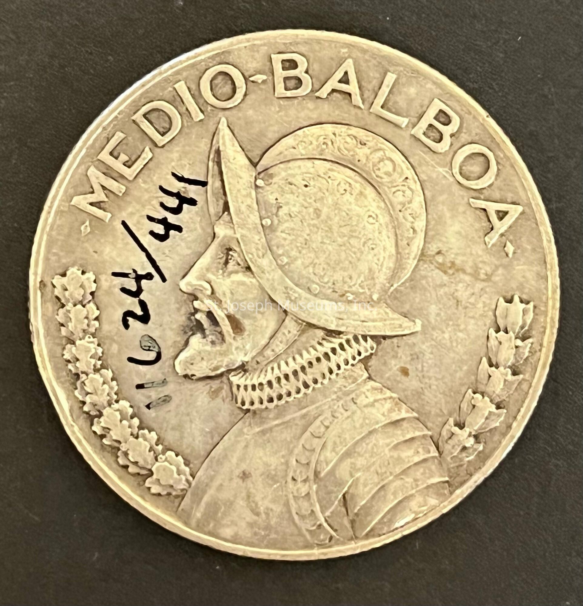 The image depicts a coin labeled "MEDIO-BALBOA," featuring the profile of a man in armor, including a detailed helmet with a patterned design, and a suit of armor, highlighted by finely engraved details, suggesting historical significance; additionally, the edge of the coin displays symbolically rich laurels on each side of the portrait, representing peace or victory, while handwritten numbers, possibly indicating a date or code, are inscribed in black ink across the coin's face, specifically over one of the laurel branches, adding a layer of mystery or notation to the otherwise historical artifact, which may be of interest to numismatists or historians alike, as the combination of elements reflects a certain period, likely emphasizing an era of exploration or conquest, common themes for such minted memorabilia from regions with a history of colonial influence or notable explorers who have been commemorated through such symbolic representations on currency, thus making the coin not only a piece of monetary value but also a captured story of human endeavor.
