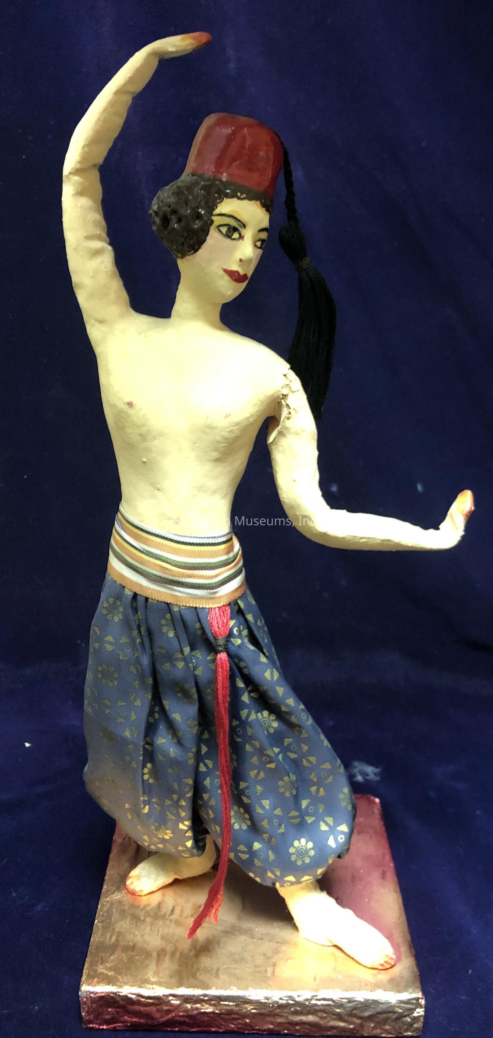 The image depicts a sculpture of a dancer in an elegant pose, wearing a red fez with a black tassel, which contrasts with short curly black hair. The figure's arms are gracefully positioned, one arm lifted above the head and the other stretched out to the side, suggesting a dance movement. The dancer's expressive face is adorned with makeup, featuring boldly defined eyes and red lips. The attire consists of loose, patterned blue pants adorned with golden floral motifs, held up by a colorful striped waistband with a red tassel hanging down. The sculpture stands on a reflective square base with a gold finish, set against a deep blue fabric background, emphasizing the figure's vibrant colors and dynamic posture.