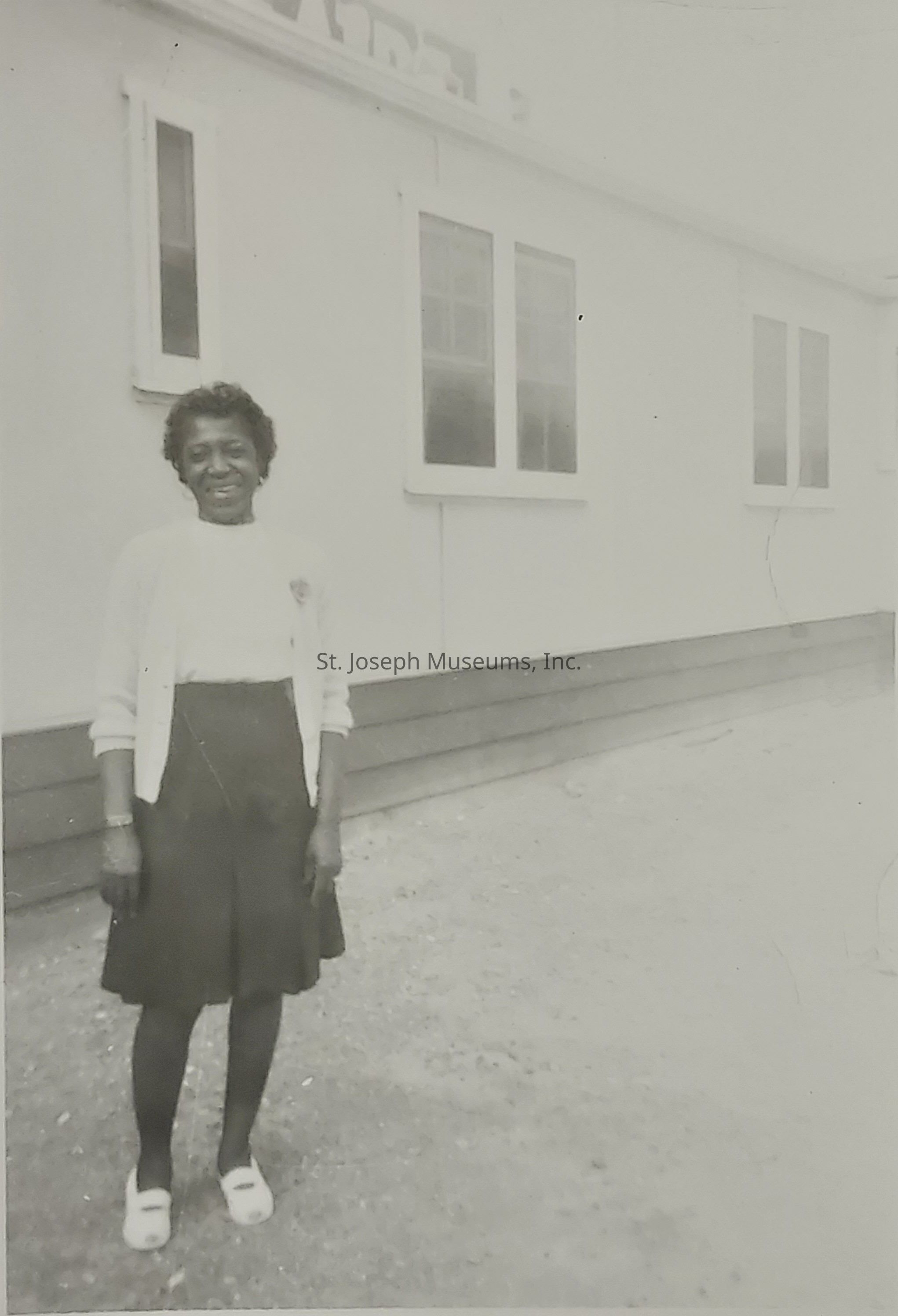 This black and white photograph captures a woman standing outdoors in front of a building, with a gravel surface underfoot. The woman is smiling gently at the camera, wearing a light cardigan over a dark skirt, dark stockings, and white open-toed shoes. Her short hair is styled neatly, and she stands with her hands at her sides, exuding an aura of calm and grace. The building behind her features several rectangular windows and horizontal siding, suggesting a possible residential or institutional setting. Despite the monochrome nature of the image, her warmth is palpable, creating a nostalgic and reflective atmosphere that invites viewers to ponder the story behind the moment captured in time.