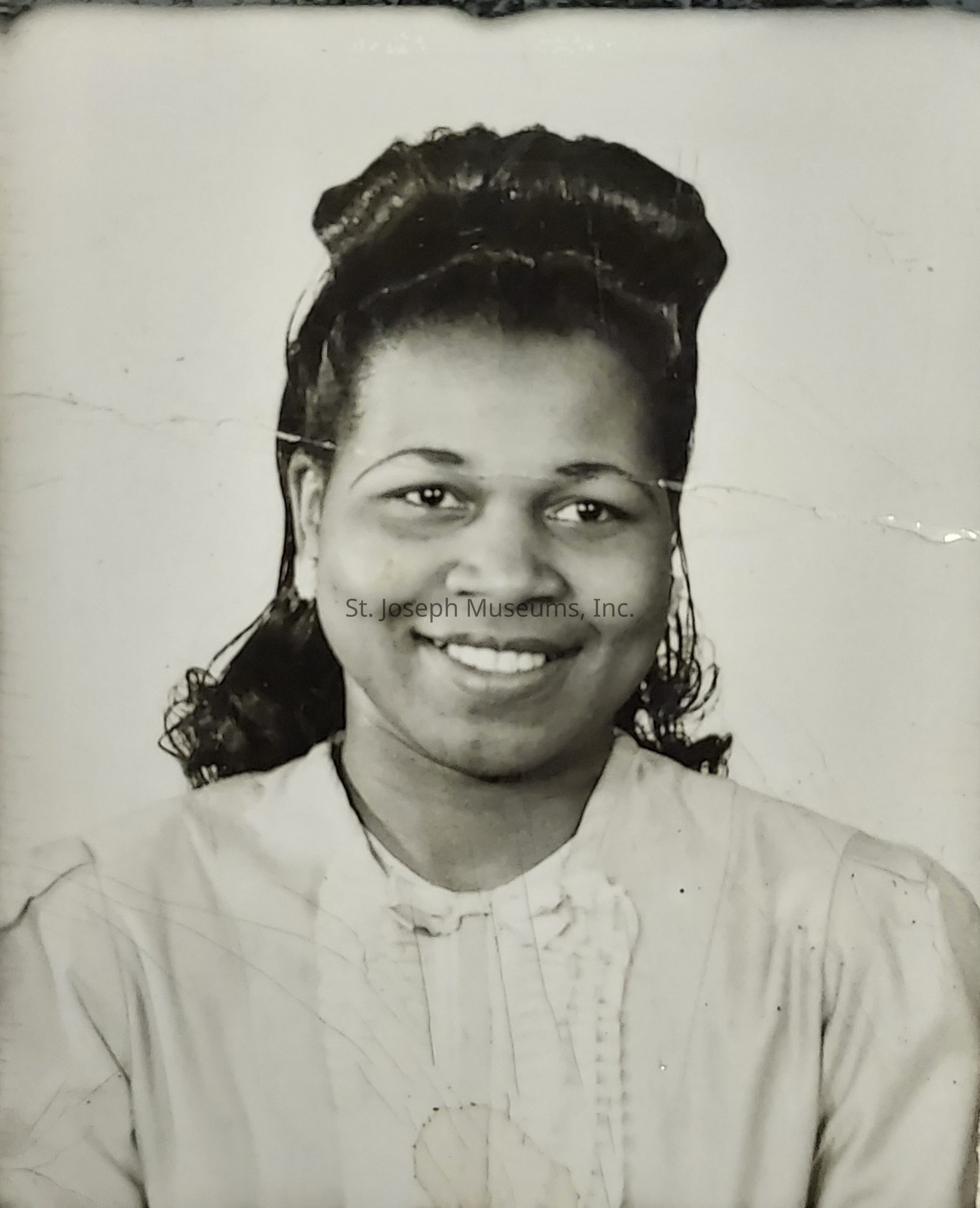 The black and white photograph features a woman smiling warmly at the camera, exuding a sense of warmth and friendliness. Her hair is styled elegantly, pulled back with volume on top, giving her a vintage look that suggests the era the photograph was taken in. She is dressed in a blouse with subtle details around the neckline, adding a touch of formality to her appearance. The image has a timeless quality, with slight creases and wear visible, hinting at its age and cherished history. Her expression is inviting and approachable, embodying a moment captured with genuine emotion.