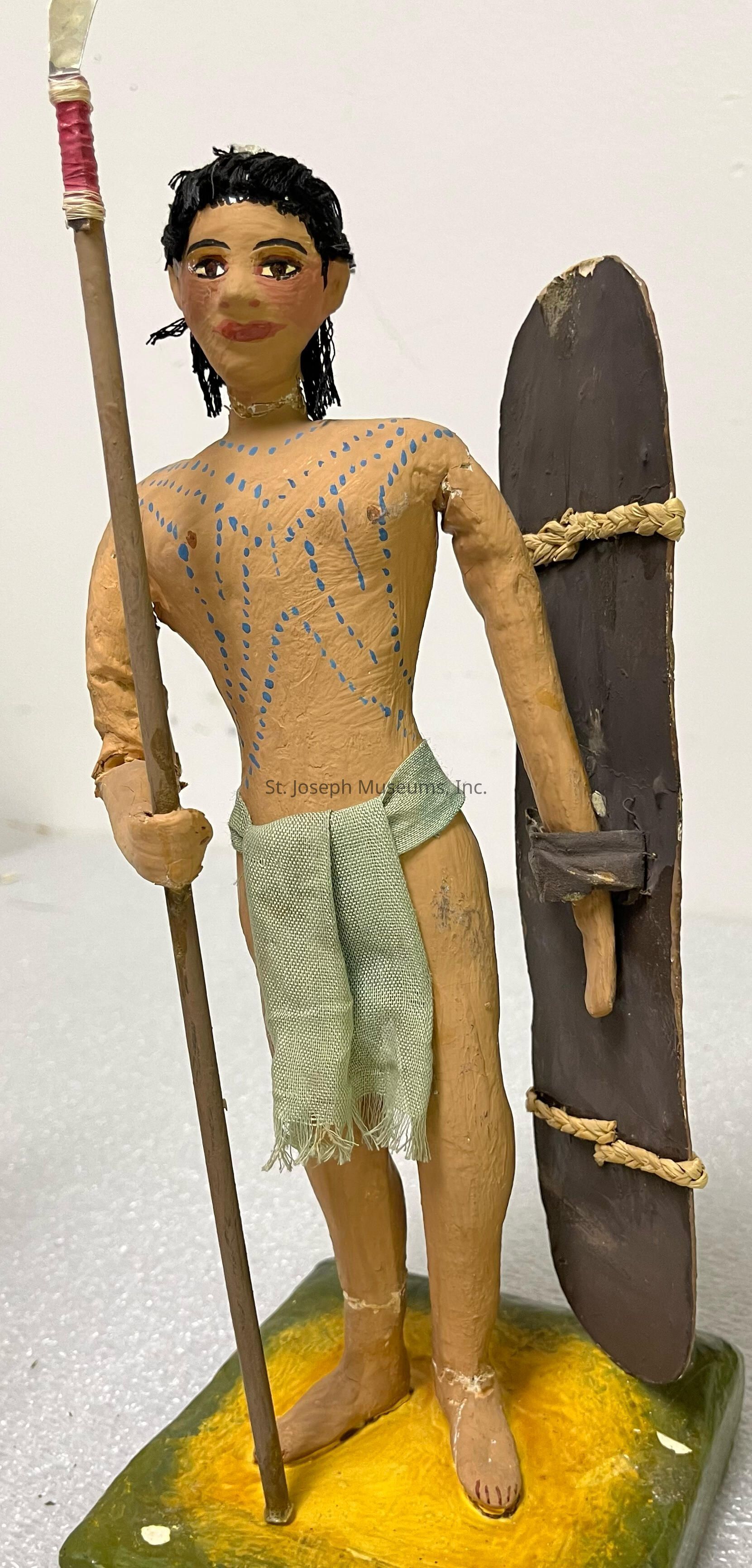 The image depicts a handcrafted figurine of a warrior, skillfully molded with a tan complexion and adorned with intricate blue patterns on the torso, giving an impression of traditional body paint. The warrior's facial features are detailed, with dark eyes and hair, exuding a stoic and determined expression. In the left hand, the figure holds a long spear with a pointed tip, wrapped near the top with red material, suggesting readiness for battle or hunting. The right arm is holding a tall, oval-shaped shield, made of a dark, sturdy material and tied with ropes, hanging protectively at the side. The attire consists of a simple, light green fabric draped as a loincloth, highlighting a minimalistic yet functional style suitable for movement and agility. The base resembles a patch of earthy ground, painted in hues of green and yellow, suggesting a natural environment. The overall craftsmanship and detail imbue the figure with a sense of cultural authenticity and historical representation.
