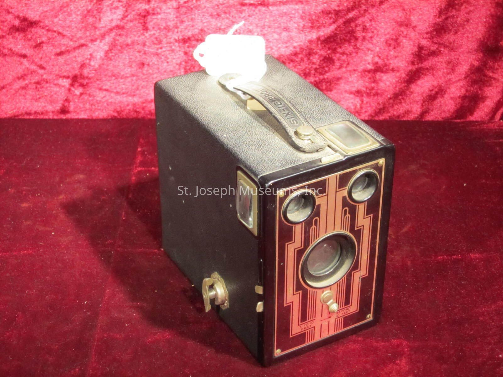 The camera has a rectangular shape with a textured black exterior and a handle on top. The front panel is designed with an intricate geometric pattern in red and gold tones, featuring three circular openings for the lenses and viewfinder. The edges of the camera are adorned with metallic accents, giving it an elegant and classic appearance. The overall design reflects an early 20th-century aesthetic, capturing the essence of antique photographic equipment with its retro charm and distinctive artistic style.