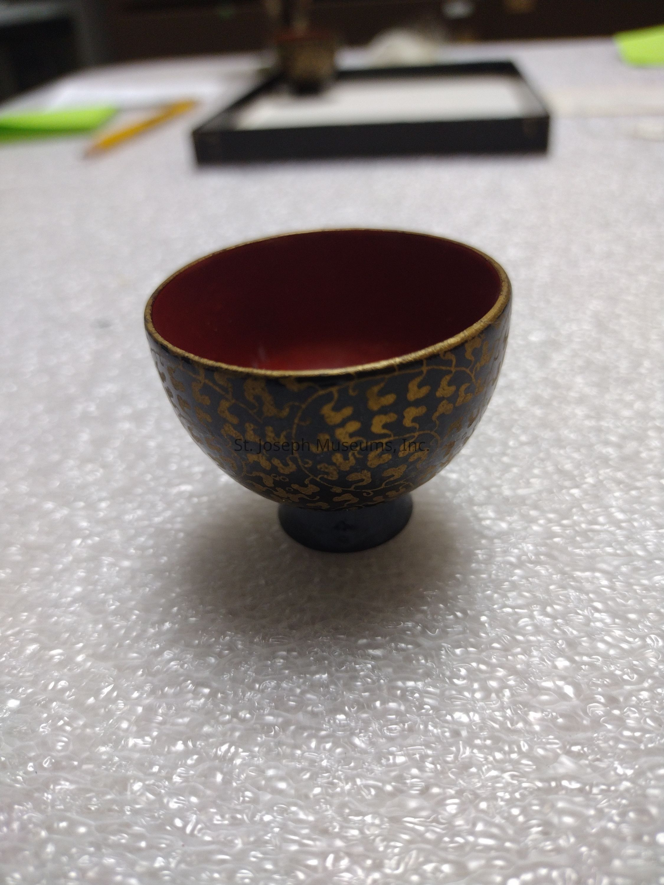 The image showcases a small, ornate cup with an elegantly rounded shape, featuring a deep red interior contrasted by a dark exterior adorned with intricate golden patterns reminiscent of traditional craftsmanship. The cup is placed on a surface covered in protective bubble wrap, hinting at careful handling or storage, possibly indicating the item’s value or fragility. In the background, a pencil and various objects are visible, slightly out of focus, resting on a tabletop, suggesting a casual or workshop setting. The overall atmosphere conveys a sense of careful artistry and attention to detail, as the design elements reflect a blend of aesthetic appeal and cultural significance, enhancing the visual allure of the cup itself.