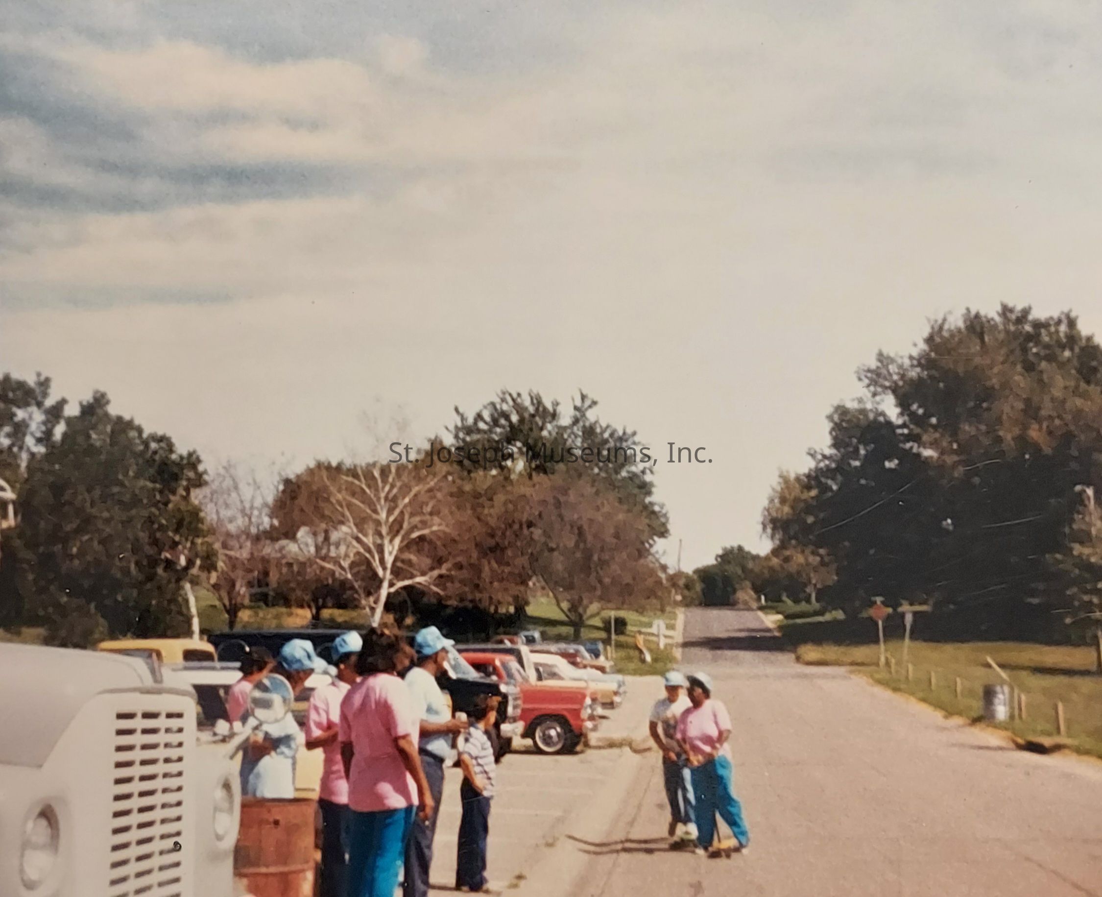The image shows a group of people gathered around a large vehicle and a barrel on the side of a road, with a backdrop of trees under a partly cloudy sky. The individuals are wearing bright blue pants and shirts, some with matching caps, perhaps indicating they are part of a team or group activity. A few children are present, and there are several parked colorful vehicles, including red and blue trucks, suggesting a possible community event or gathering. The road stretches into the distance with green grass and more trees lining the sides, creating a tranquil suburban atmosphere.