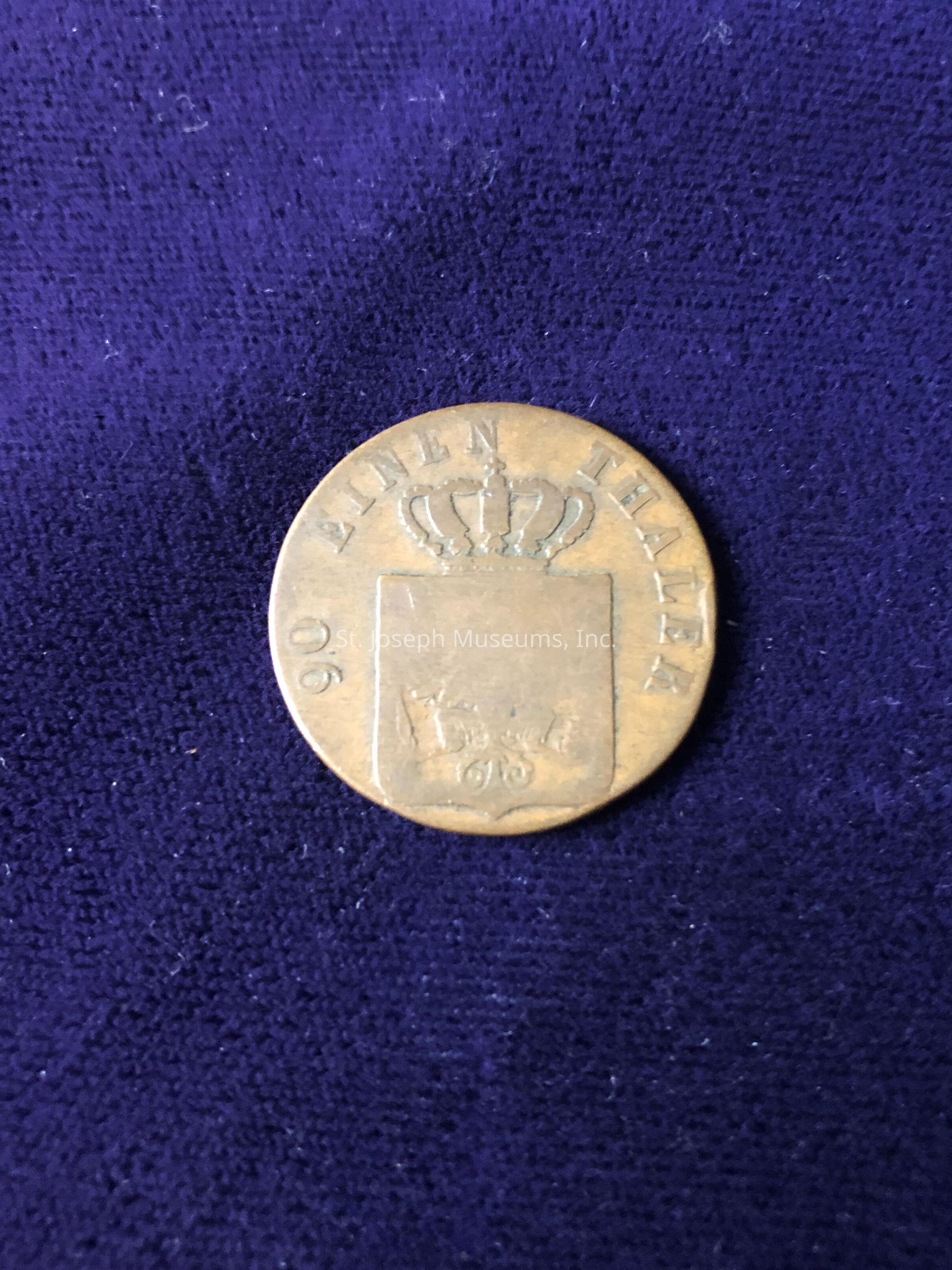The coin, with a noticeable patina indicating its age, features the text "EINEN THALER" along the edge. At the center of the coin is a design containing a crown above a shield. The number "90" appears on the left, and there seems to be a "95" or similar inscription on the bottom of the shield, possibly indicating a date or value. The purple background provides a rich contrast that highlights the coin's metallic surface and intricate details. The coin appears to have historical significance, possibly originating from a region where the Thaler was used as currency.