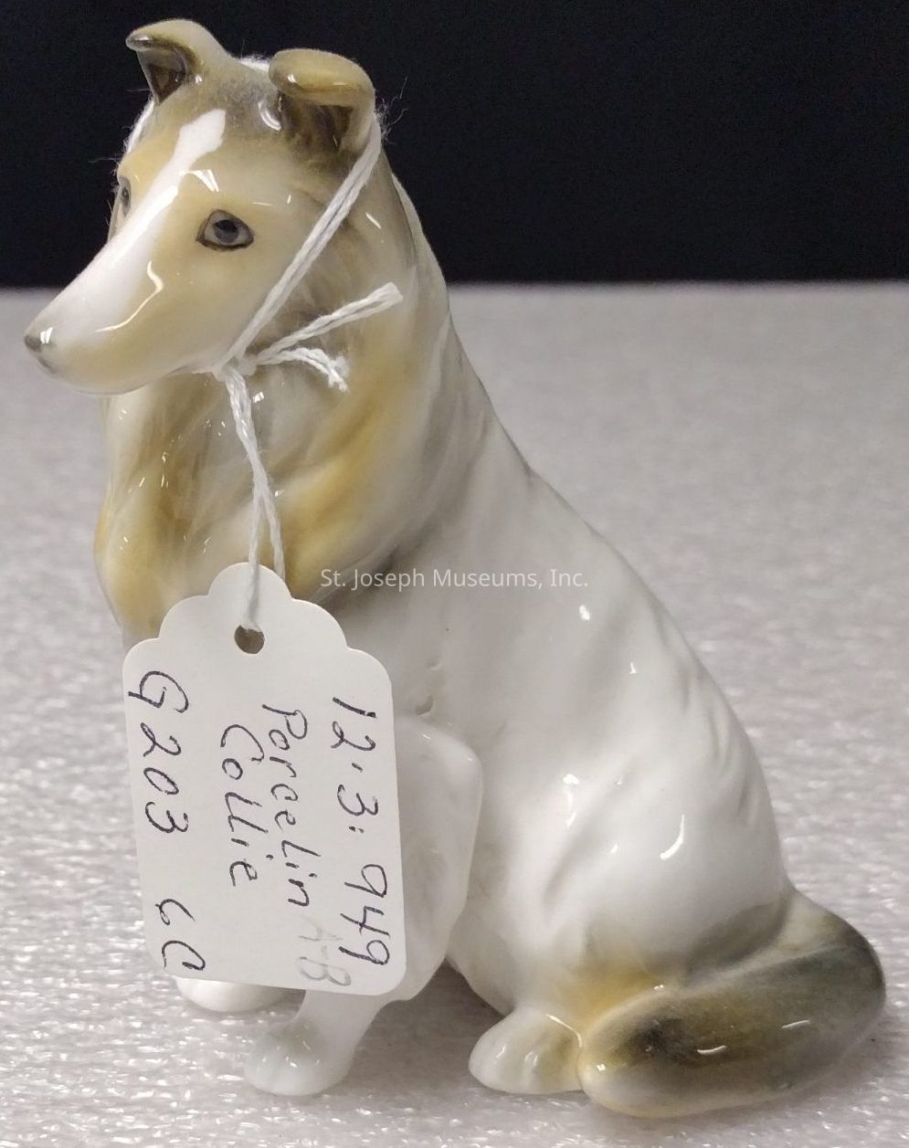 The image depicts a porcelain figurine of a Collie dog, beautifully crafted with attention to detail, capturing the likeness of the breed's characteristic features such as its pointed ears and long coat. The figurine is primarily painted in shades of white and tan, giving it a realistic appearance. A small tag attached to the figurine reads "Porcelain AB Collie" along with additional information such as a date, "12.3.949," and some letters and numbers, "G203 CC." The figurine is placed on a light-textured surface, and its glossy finish reflects the light, enhancing its delicate craftsmanship. This piece likely serves as a collectible item, appreciated for both its artistic value and its representation of the beloved Collie breed.