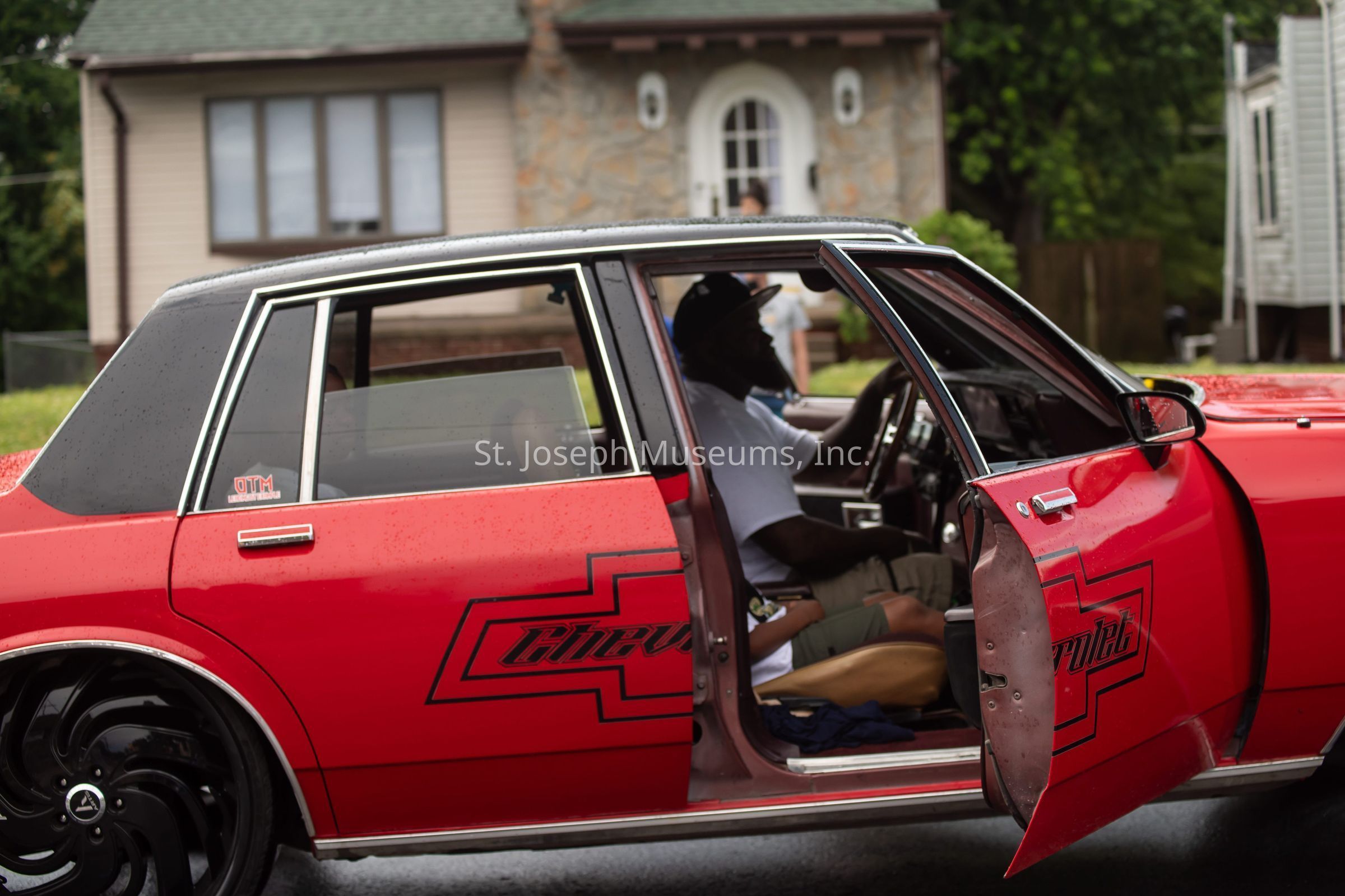 The image showcases a vibrant red Chevrolet sedan parked on a residential street, with its front door open to reveal a man sitting inside, dressed in casual attire with a white t-shirt and cap, appearing relaxed as he occupies the driver’s seat. The car, notable for its retro design, features bold, custom black graphics on the door, including a stylized logo, and is equipped with large, glossy black rims that add a distinctive flair to the vehicle's appearance. Raindrops are visible on the car's surface, suggesting a recent or ongoing light rain, while in the background, a quaint house with a stone facade and a well-maintained lawn provides a suburban setting, further emphasizing the leisurely street scene. The overall composition of the image captures the fusion of automotive enthusiasm and everyday life, where the classic car becomes a central character in the narrative, set against the backdrop of a typical neighborhood.