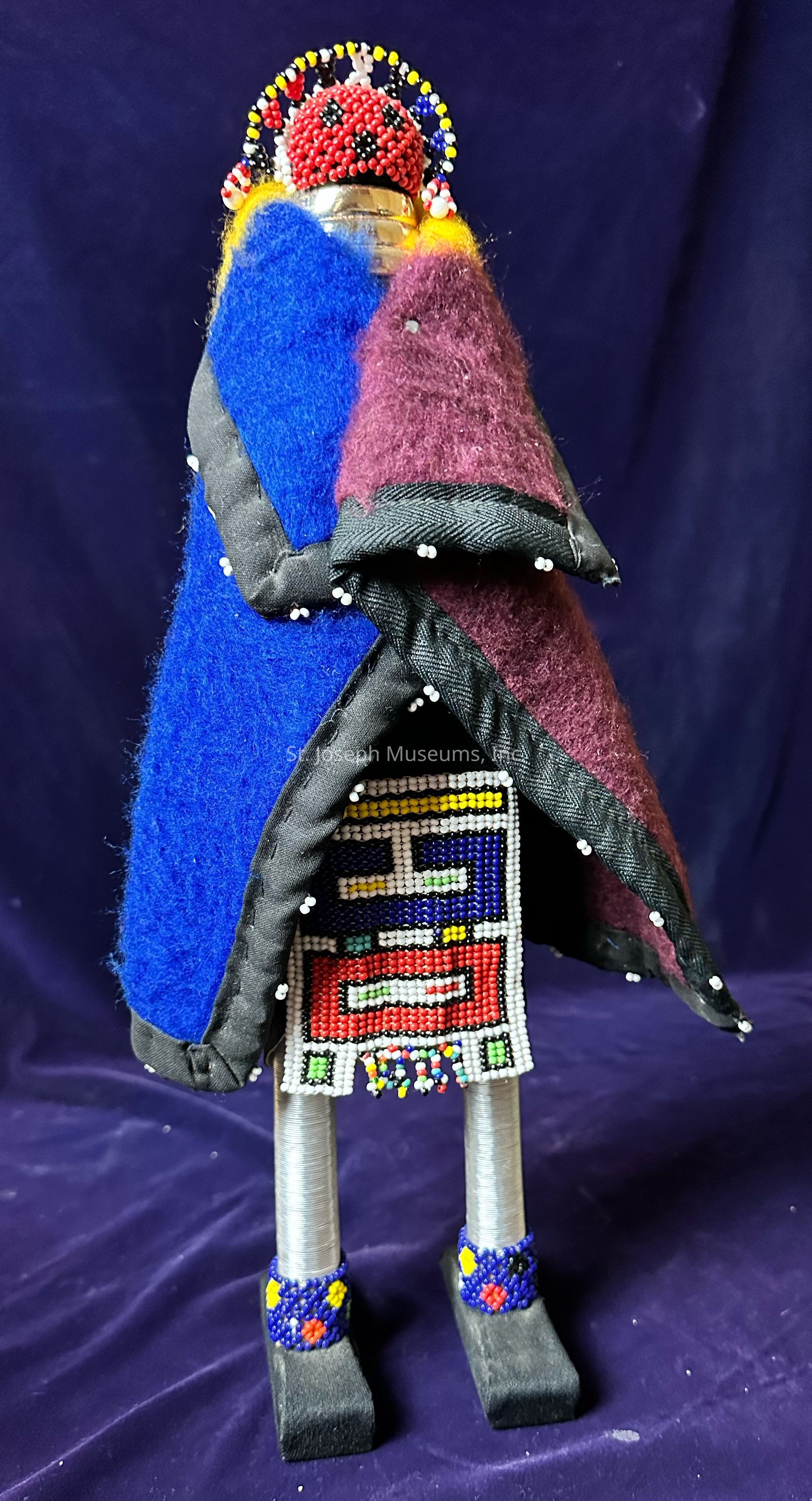 The image depicts a handcrafted doll adorned with intricate beadwork and vibrant textiles against a deep purple background. The doll's head, made of beads in red, black, and white, creates a striking pattern, while its body is enveloped in colorful layers of fabric in shades of blue and maroon, with a black border decorated with small white beads. The front of the doll features a detailed beaded design in various colors, including blue, red, yellow, and green, creating a visually captivating centerpiece. The lower part of the doll is composed of coiled metallic wires, forming its legs, which are embellished with beaded anklets in blue, yellow, and red, resting on square black bases that resemble shoes. This artistic piece combines an array of textures and patterns, showcasing cultural craftsmanship and attention to detail, presenting an eye-catching display of traditional artistry and vivid color contrasts.