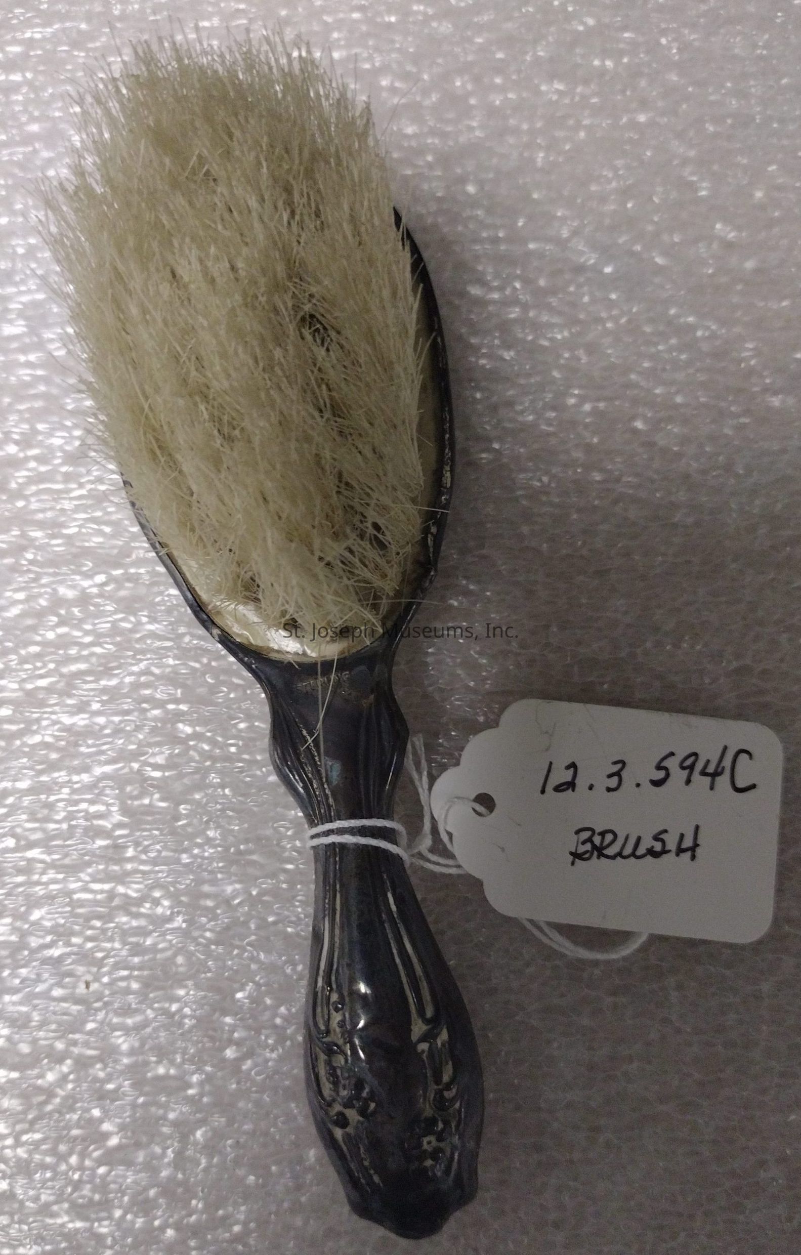 The image shows an elegant, vintage hairbrush featuring a decorative dark handle with ornate designs, complemented by a cluster of light-colored, dense bristles at the top. The brush is positioned against a softly textured, light background, which highlights its details. Attached to the brush's handle with a piece of white string is a small, white, scallop-edged tag that is inscribed with the numbers and letters "12.3.594C" and the word "BRUSH" in handwritten, bold lettering, suggesting categorization or archival purposes. This tag adds an element of historical or collectible significance to the object, indicating it may be part of a collection or exhibit. The overall composition of the image evokes a sense of nostalgia, craftsmanship, and an appreciation for the artistry and functionality of bygone eras, offering a glimpse into the design aesthetics and craftsmanship valued in past generations.