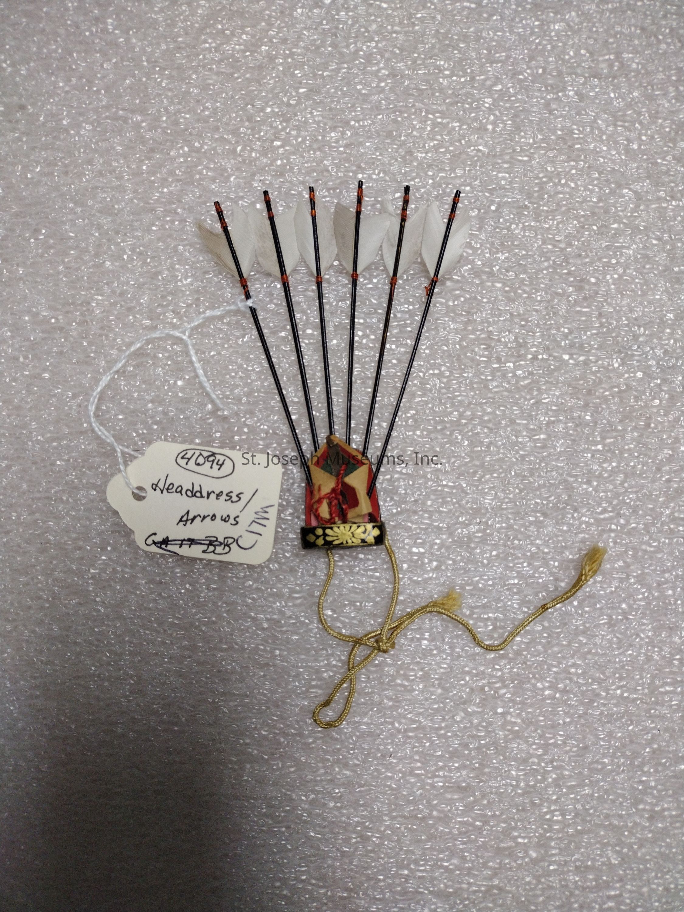The image features a decorative headdress made of six miniature arrows, each with a black shaft, red and black colored tips, and white fletching. These arrows are arranged in a fan-like pattern, emerging from an ornate base adorned with red and gold floral motifs. A delicate golden string is attached to the base, coiled neatly below. The headdress is placed on a textured, bubble-wrap-like surface, providing a contrasting backdrop. Attached to the headdress is a white label with handwritten text, including numbers and the words "Headdress/Arrows," which suggests some sort of cataloging system. Overall, the image highlights a unique and artistic representation of cultural or ceremonial adornment, reflecting meticulous craftsmanship and attention to detail. The combination of the traditional design elements and modern catalog tag creates an intriguing juxtaposition, enhancing the object's appeal and inviting curiosity about its origins and purpose.