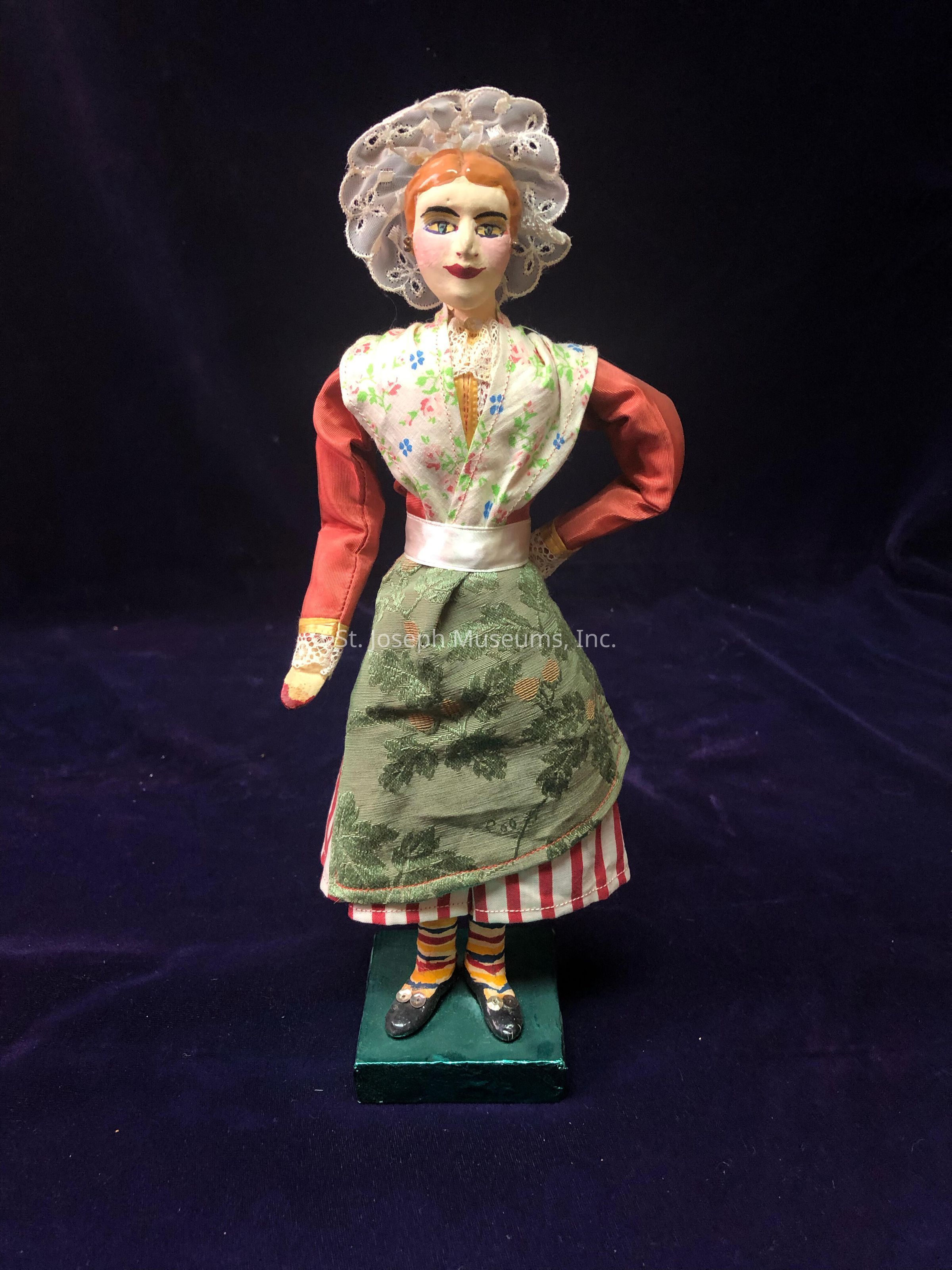 The image portrays a detailed handcrafted doll, set against a dark, velvety backdrop. The doll is dressed in a traditional outfit consisting of a vibrant red top with long sleeves and a green skirt featuring intricate floral patterns. Her ensemble is complemented by a floral apron and a sash tied at the waist. She wears striped and colorful socks paired with black shoes, adding a playful touch. Her hair is styled neatly beneath a lace-adorned bonnet, exuding an old-world charm. The doll’s expressive face, with carefully painted features and rosy cheeks, reflects meticulous craftsmanship and artistic skill. This charming figure stands confidently on a green base, showcasing both cultural heritage and artistic creativity.