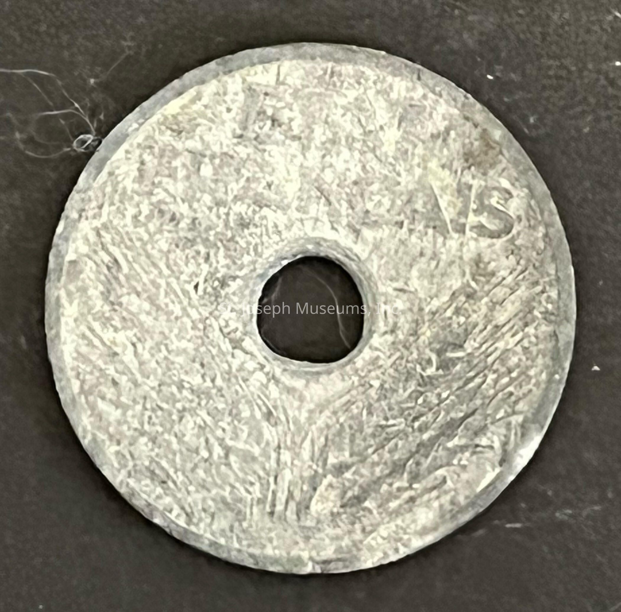 The image depicts an aged, circular, metallic object resembling a coin with a noticeable hole in its center. The surface of the coin is rough and weathered, suggesting it is old or made to appear distressed, lending it a rustic and historical aura. Indistinct markings or engravings are present on the surface, with some characters visible, although they are not entirely legible due to the wear. The raised edge of the coin is slightly rounded, adding dimension to its appearance. Loose strands or fibers near the edge indicate the coin may have been in contact with other materials.