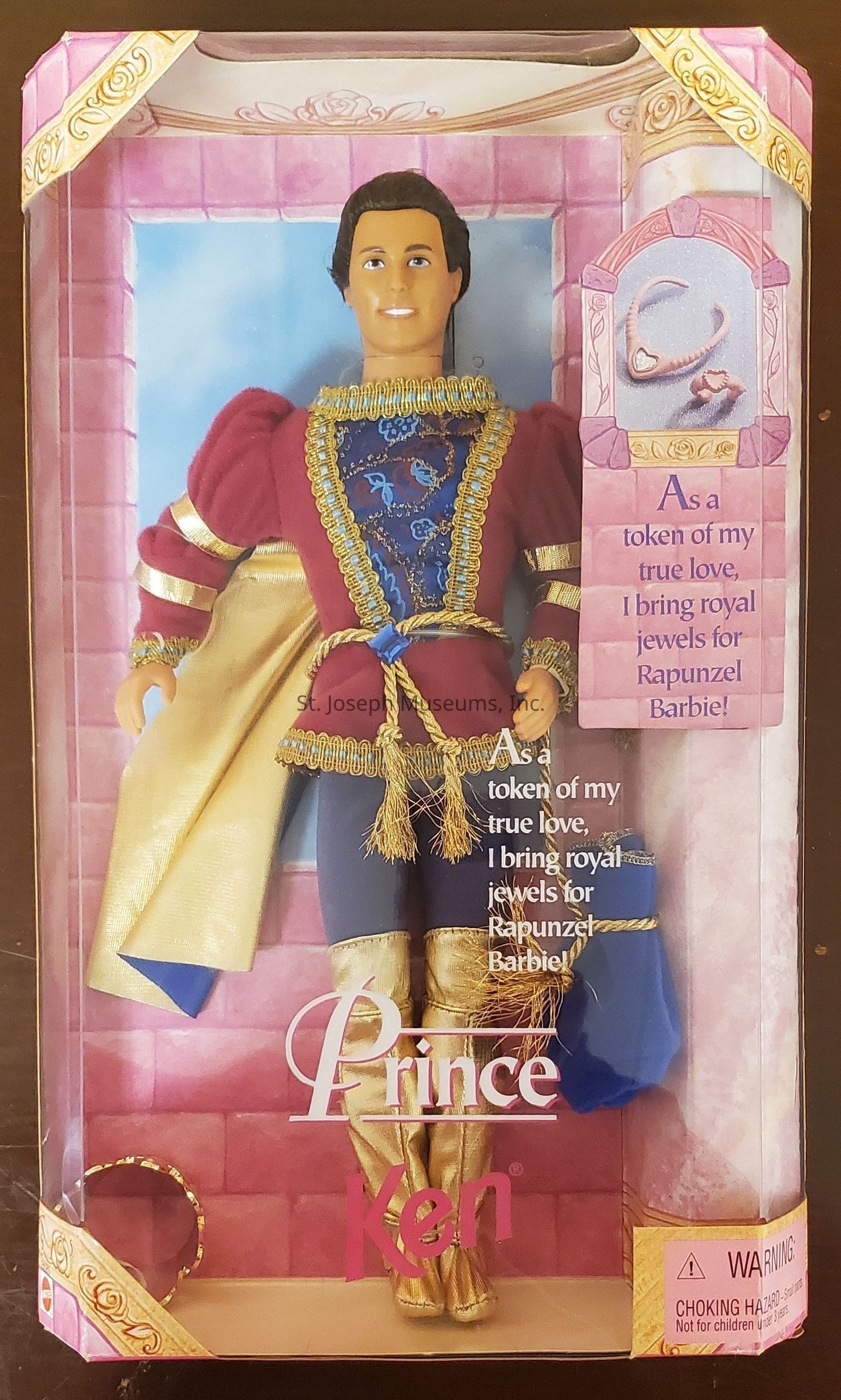 The image depicts a boxed Prince Ken doll designed to complement the Rapunzel Barbie collection. The doll is dressed in an elaborate and regal outfit that includes a deep red tunic with golden and blue embroidery, paired with royal blue trousers. His attire is further enhanced by gold embellishments, including a golden sash and gold boots that add to the royal theme. The background inside the box presents a pink castle setting, reinforcing the fairy tale ambiance. The packaging has decorative gold patterns and contains text that reads, "As a token of my true love, I bring royal jewels for Rapunzel Barbie!" Additionally, there's a visible warning for choking hazards, indicating the toy's suitability for children aged three years and older.