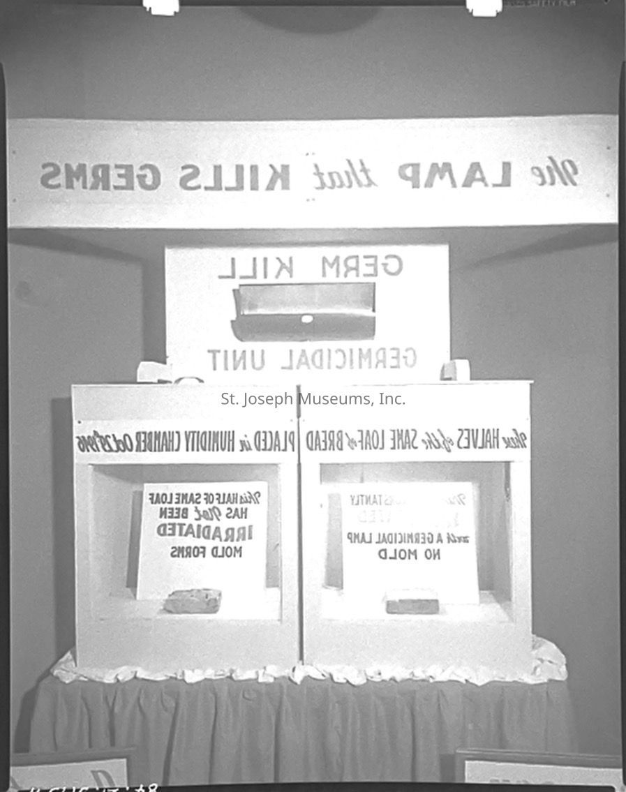 A black-and-white photograph from the St. Joseph Light and Power Company showcasing a window display at 6th & Francis Street featuring germ-killing lamps, with text highlighting their sanitizing properties. The display includes two sections comparing irradiated and non-irradiated bread, showcasing the effectiveness of the lamps in killing germs.