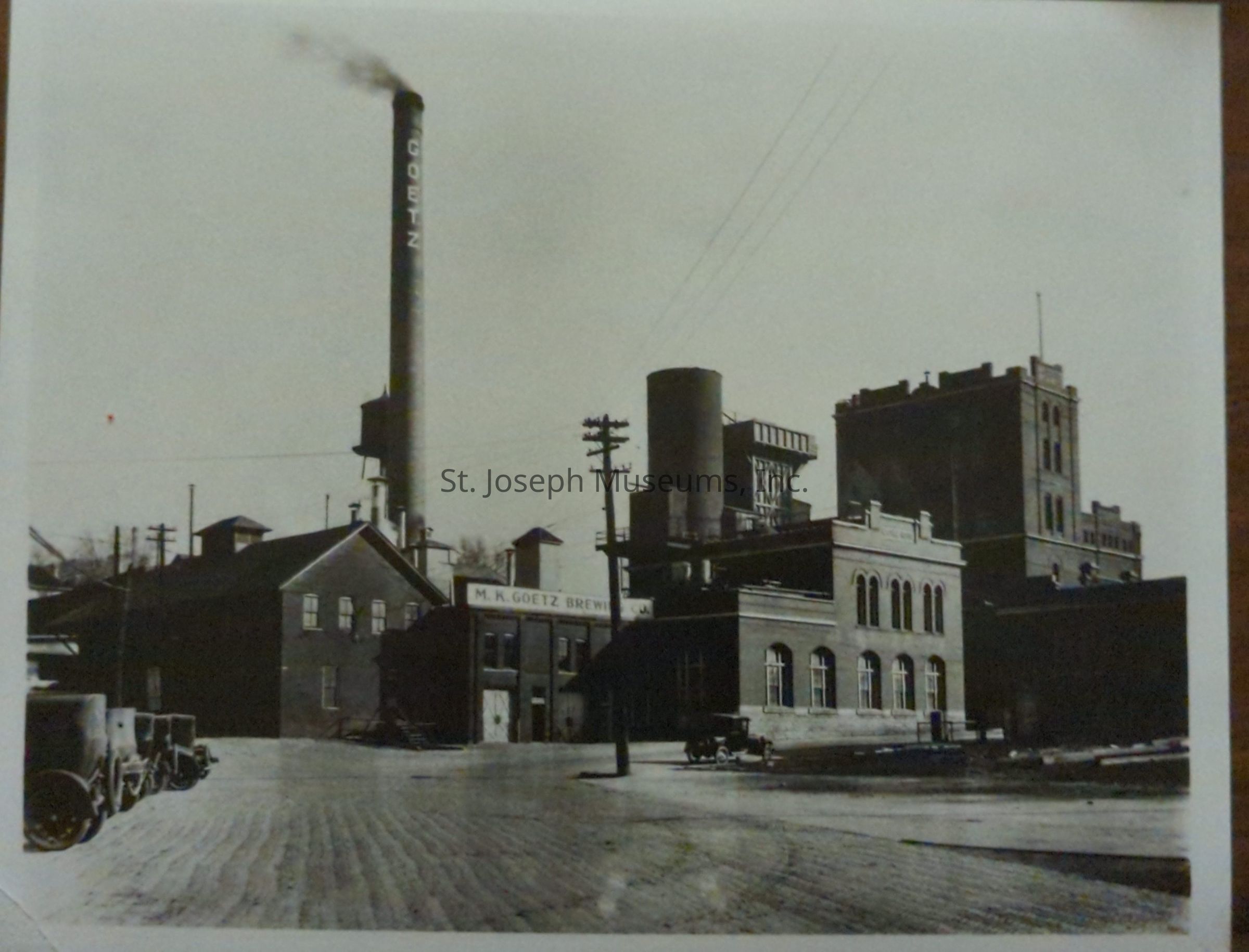 Black-and-white photograph depicting the M.K. Goetz Brewing Company located at 601 Albemarle in St. Joseph, Missouri. The industrial site features several large brick buildings and a prominent smokestack emitting smoke, with the word "GOETZ" visible on the stack. Vintage cars are parked on the cobblestone street.