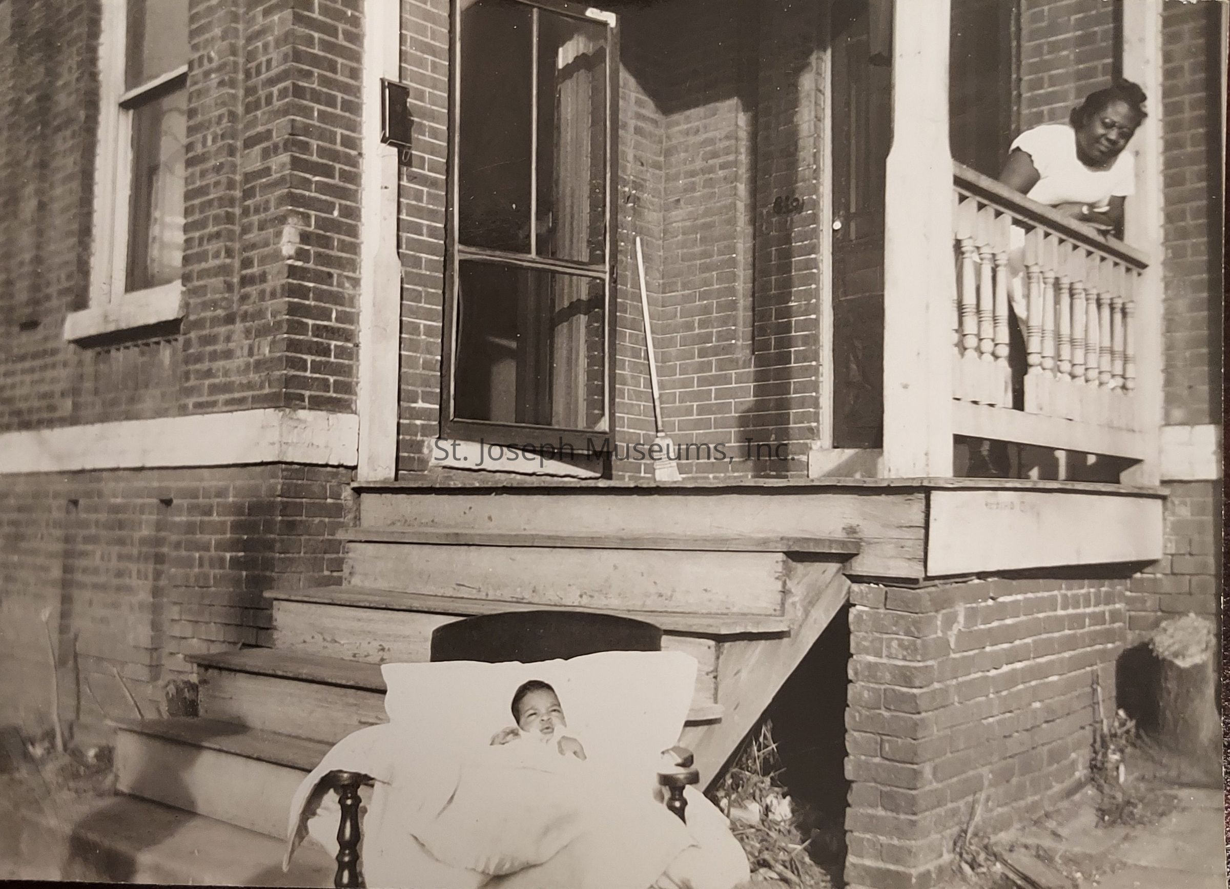 The black and white image captures a serene moment outside a brick house with an inviting porch. A woman leans over the porch railing, smiling warmly as she gazes at an infant cozily nestled in blankets on a chair at the bottom of the wooden steps. The porch features architectural details like a screen door and a spindle railing. A broom stands against the wall, suggesting a home recently tidied. The infant, lying peacefully, adds a tender element to the scene, representing new life and family connection. The play of light and shadows on the bricks creates a soothing, timeless ambiance. In the background, the afternoon sun casts gentle shadows, highlighting the texture of the bricks and the smoothness of the wood. The quiet charm of the scene evokes a sense of warmth and simplicity, reminiscent of early to mid-20th century life. The image tells a story of familial affection and everyday moments, beautifully captured in a single frame, hinting at the rich history embedded in this domestic setting.