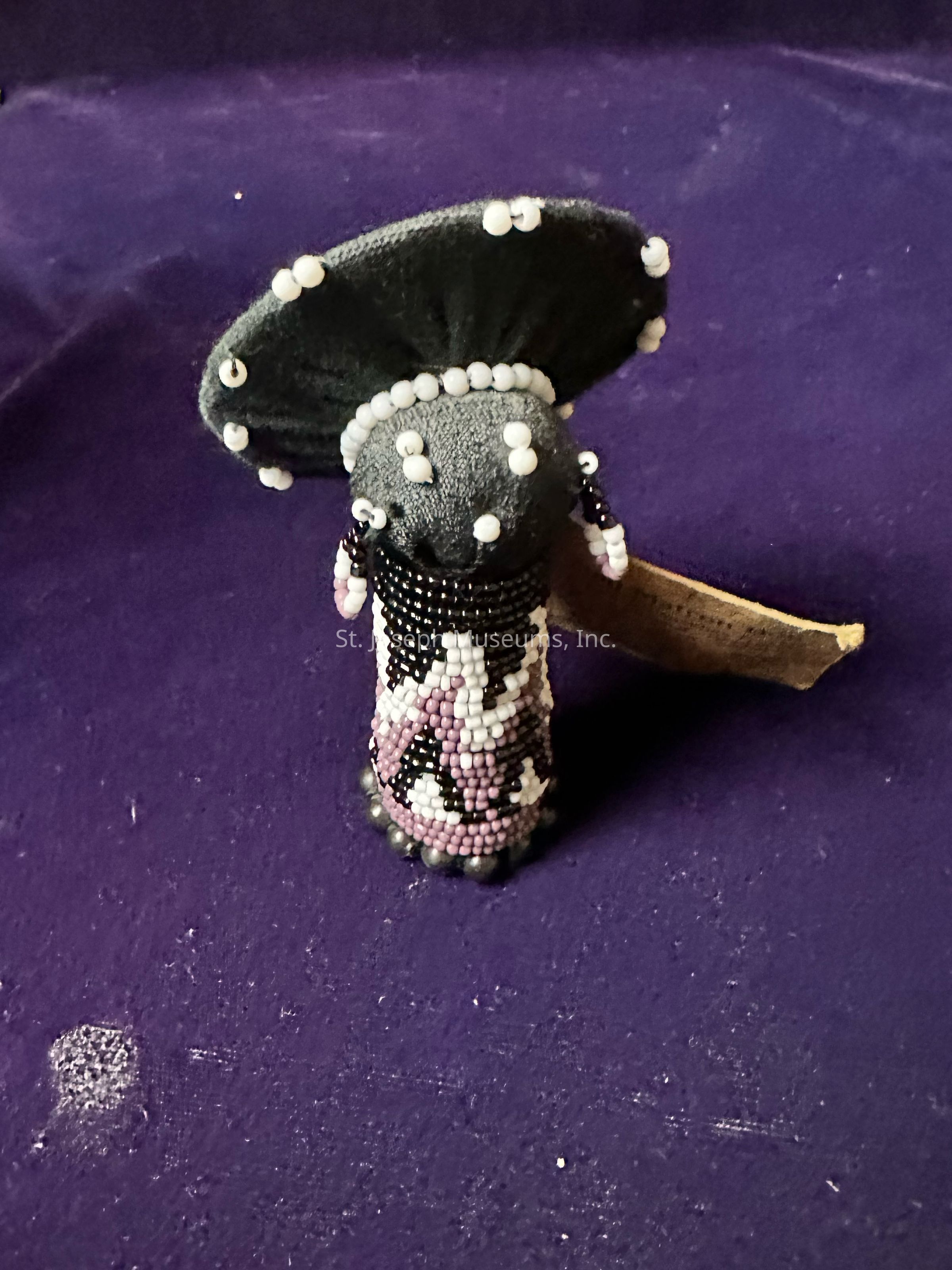 The image displays a creatively crafted object resembling a mushroom with a broad, black fabric cap adorned with small white beads that are evenly spaced around the edge, creating a striking contrast against the dark textile. Its stem, covered in a mesmerizing pattern of numerous small beads in shades of black, white, and pink, suggests intricate artistry and careful attention to detail. The object stands on a purple fabric surface, which serves as a complementary background to enhance its overall aesthetic. Alongside the mushroom, there appears to be a small piece of aged, worn paper, partially visible, adding an element of curiosity and context to the scene. The mushroom's distinct materials and vibrant design reflect a fusion of craft and creativity, capturing the viewer's attention and inviting admiration for the work's meticulous beadwork and artistic vision.
