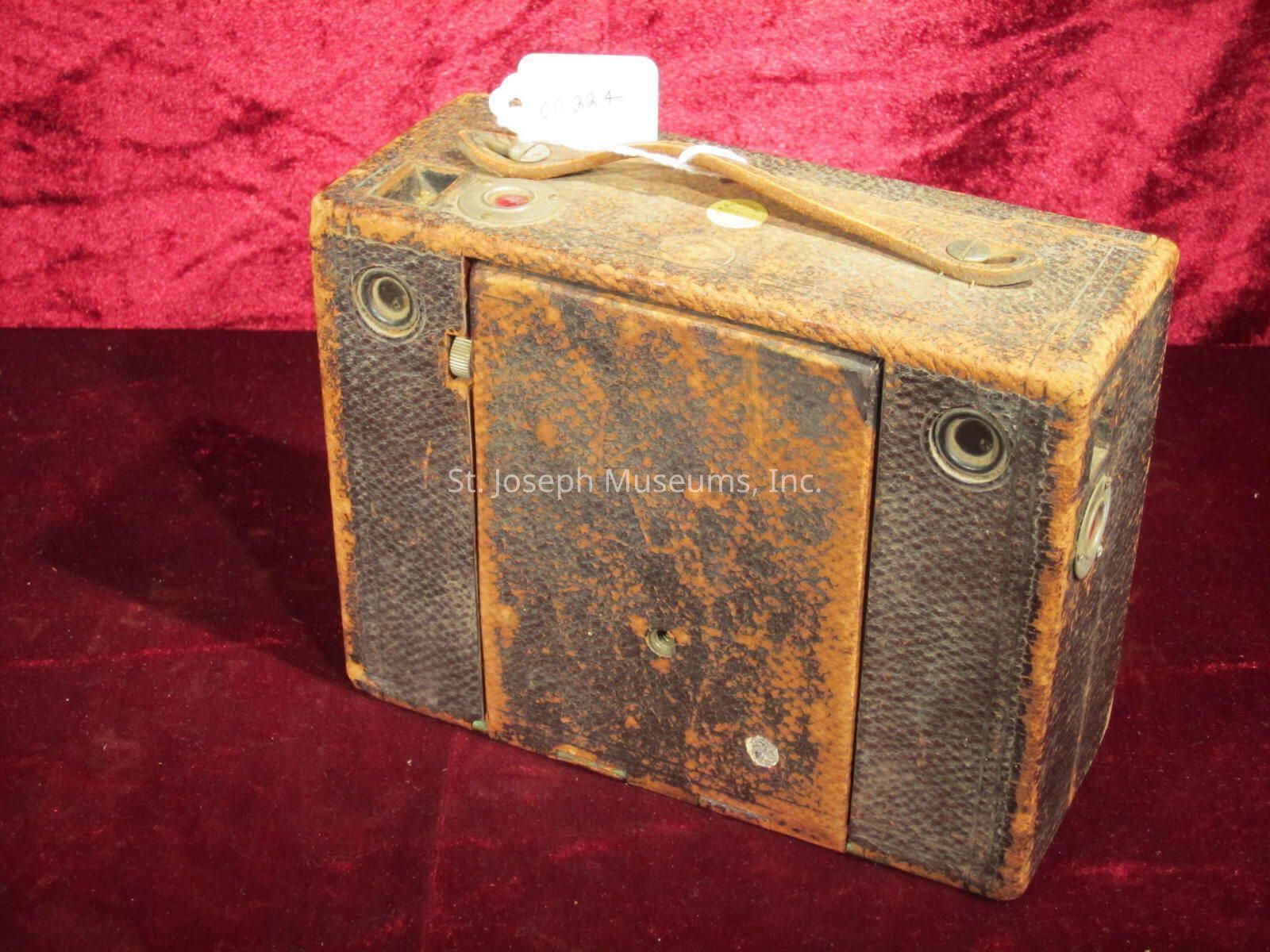The image displays a vintage box camera, characterized by its rectangular shape and worn, textured exterior. The camera appears to be made from a sturdy material, possibly leather or a similar fabric, which shows signs of aging, with visible scuffs and a rich, darkened patina indicative of its historical nature. It features a simple design with minimal buttons or controls, suggesting basic functionality. There is a small, circular lens centered on the front, flanked by metal components, which are likely part of the camera's operation mechanism. On the top, a small leather strap hints at its portability, allowing easy transport when in use. The camera sits against a plush, deep-red velvet backdrop, adding a contrasting touch that enhances its antiquarian appeal and highlights its craftsmanship.