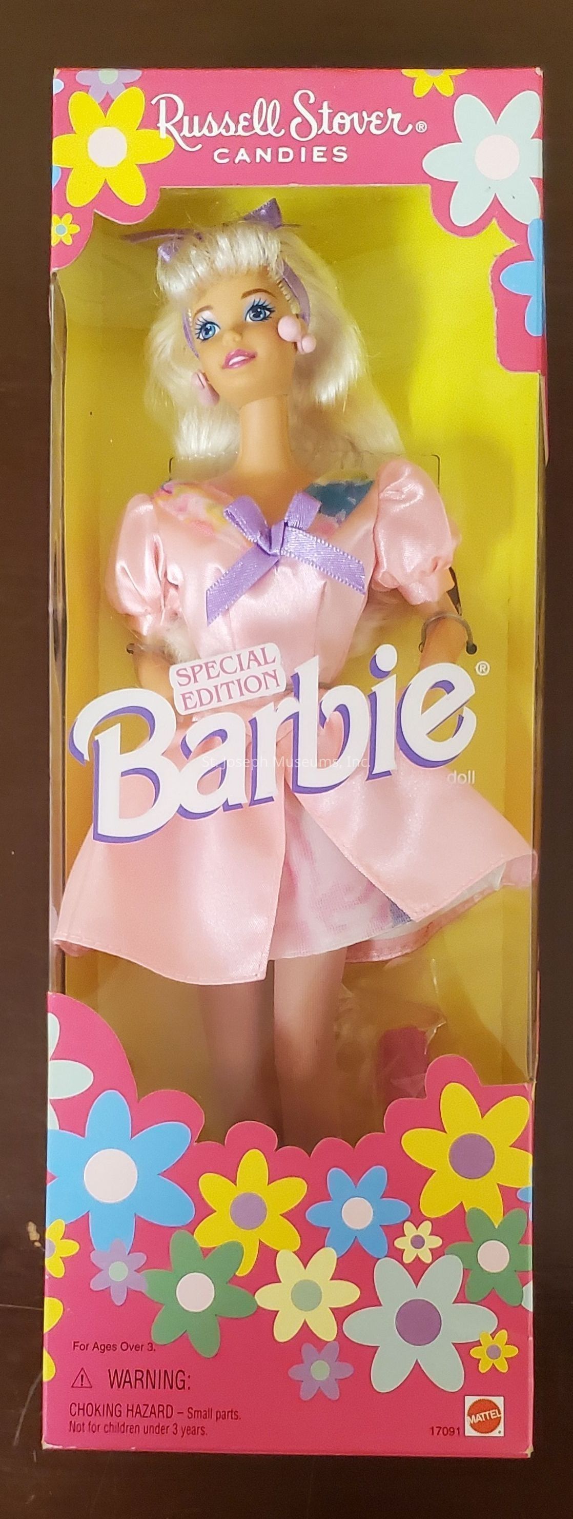 The image displays a special edition Barbie doll encased in its original packaging. The box is vibrant, featuring a floral design with pink, yellow, blue, and green flowers. At the top, there's the "Russell Stover Candies" branding, indicating a collaboration. The Barbie, visible through the clear front, is wearing a shiny pink dress with puffed sleeves adorned with a purple bow, and she has long blonde hair styled with pink accessories. The background of the box is yellow, enhancing the doll's colorful attire. Notably, it includes a warning about small parts, advising it is suitable for ages over three. The bottom right corner features the Mattel logo, confirming its authenticity. This collectible edition seems to highlight a playful and nostalgic theme, merging candy and Barbie aesthetics, likely appealing to collectors and enthusiasts who cherish unique and themed editions.