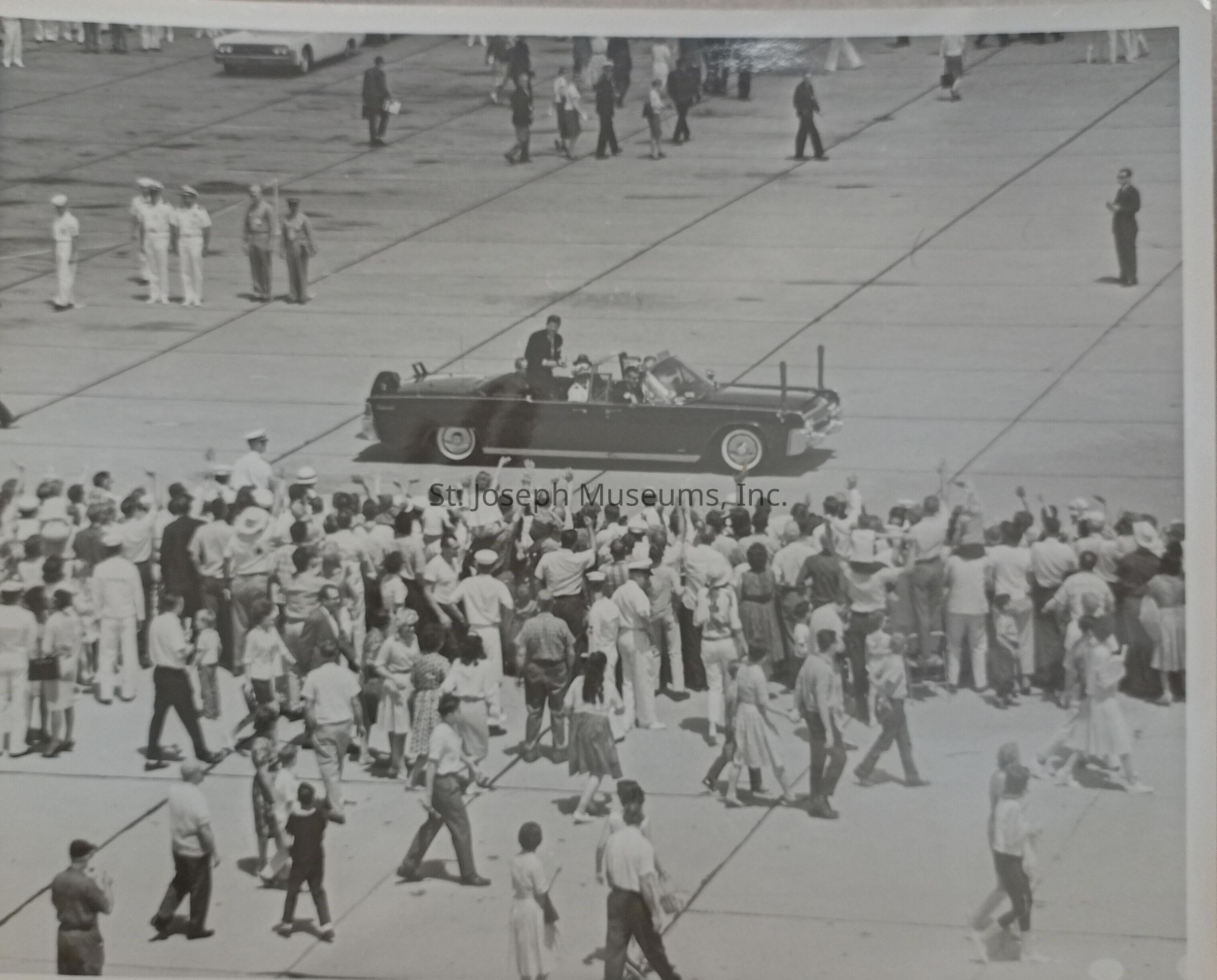 Black-and-white image showcasing John F. Kennedy on the campaign trail in a black convertible driving on the runway at Rosecrans Airport, Kansas City, surrounded by an enthusiastic crowd. The crowd, composed of men, women, and children, eagerly waves and cheers as the car passes by. Several people are visible on the tarmac, and uniformed officials stand in the background.