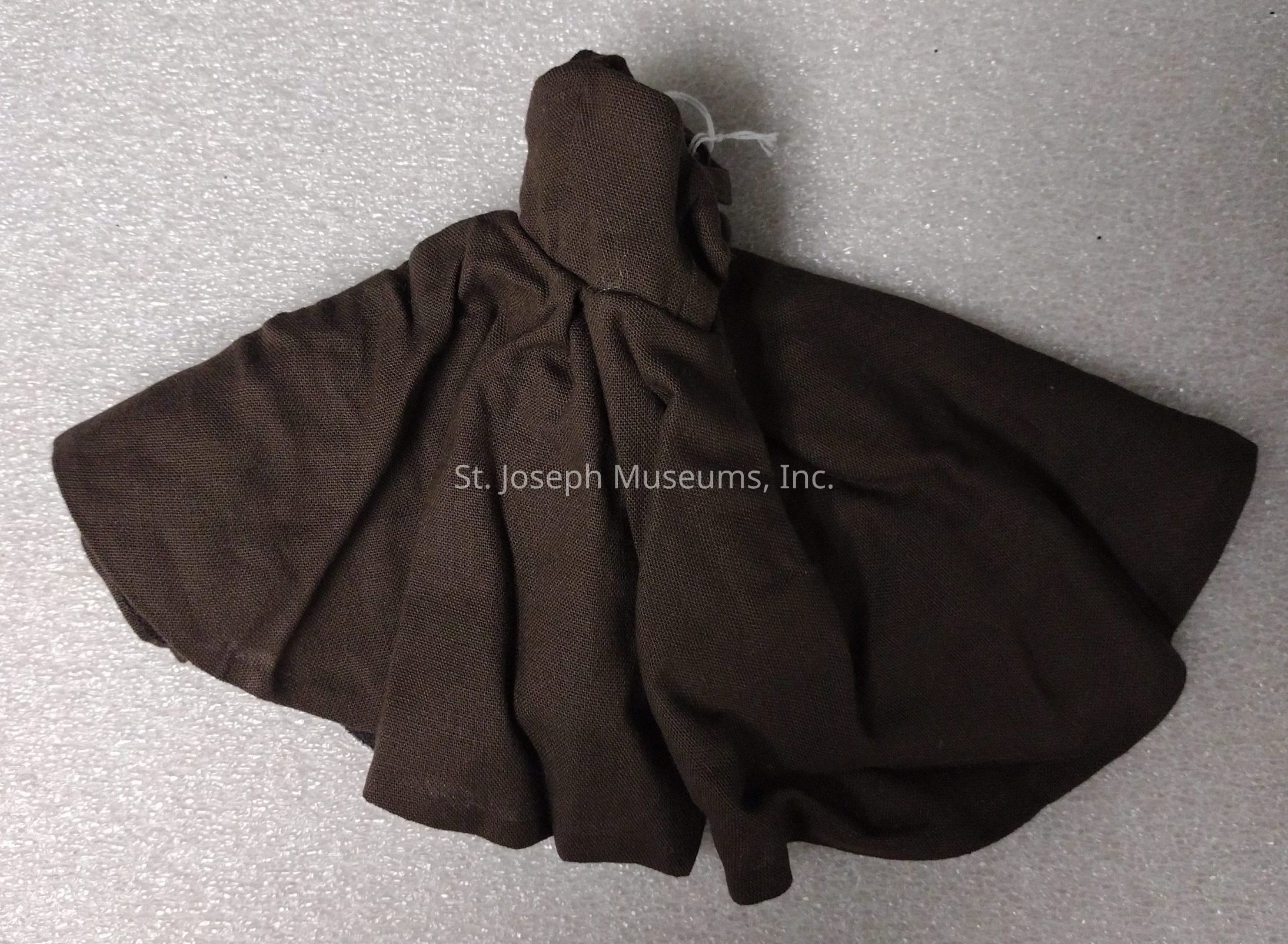 The image shows a dark brown fabric piece resembling a cloak or cape with a raised, gathered section at the top that might be intended to represent a hood. The fabric is textured and appears to be made from a thick, rugged material, possibly designed for a toy or model. It is spread out against a light, gray, textured background, highlighting the folds and draping of the material, which is arranged in such a way that it creates a sense of movement or flow, as if caught in a gentle breeze. The construction suggests simplicity, with visible stitching and unrefined edges, possibly indicating a hand-made creation or a piece designed for a specific utilitarian or artistic purpose, such as a costume, model accessory, or decorative item. This garment-like item evokes a sense of mystery or adventure, potentially conjuring images of a miniature figure enshrouded in secrecy or embarking on a hidden quest.