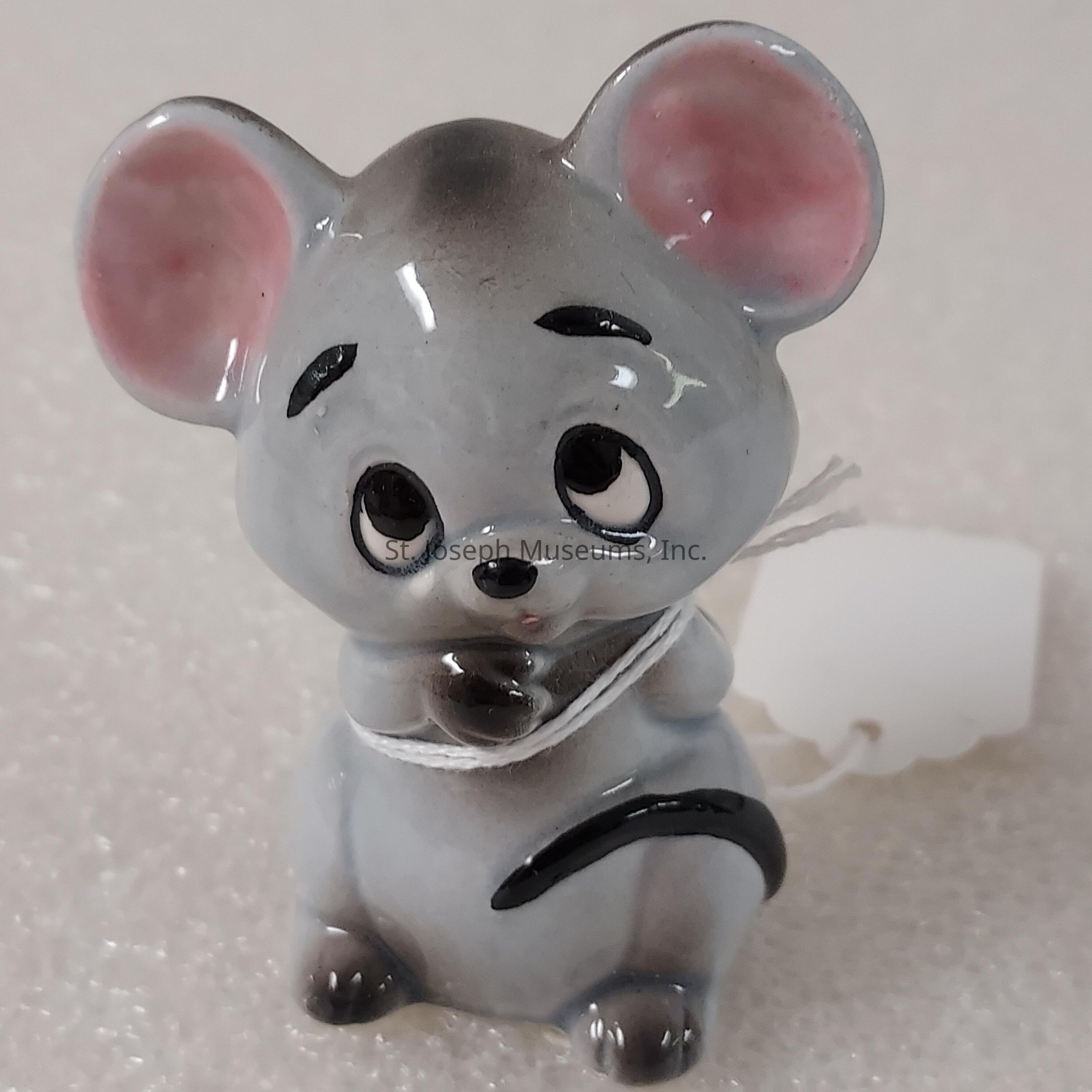 The image shows a cute, ceramic mouse figurine with large, pink inner ears and a glossy gray body. Its face features wide, expressive eyes and small, arched eyebrows, giving it an adorable, curious expression. The figure is sitting upright, paws held in front of its body, wrapped with a thin piece of white string. There is a small, black-tipped tail curving around its back. The mouse is placed on a light, speckled surface with a small, white, rectangular tag attached, suggesting it might be a collectible item or a decorative piece. The craftsmanship appears detailed and polished, highlighting its whimsical charm.
