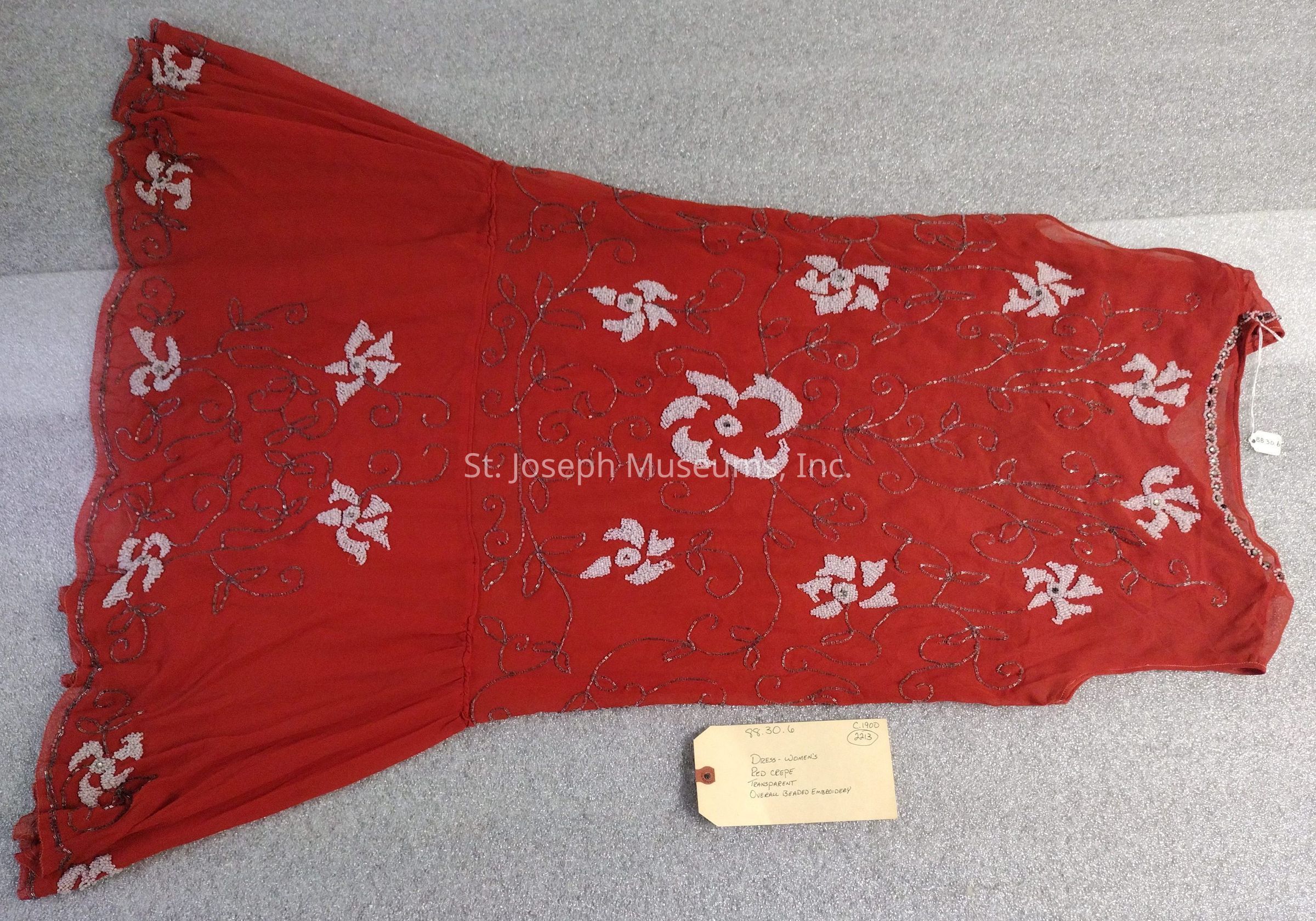 This is a knee-length, sleeveless dress made from red-colored crepe material, featuring a transparent design without lining. The dress is adorned with overall beaded embroidery in a floral pattern using white and clear beads. It includes some rhinestones placed at the neck and in the centers of the flowers.