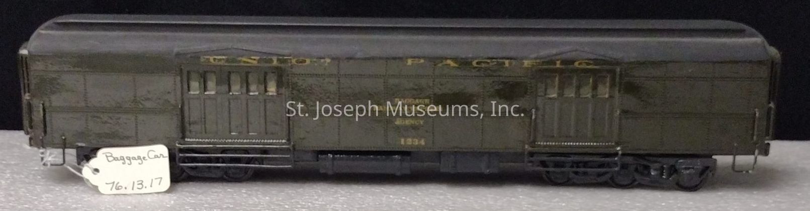 The image depicts a model train baggage car displayed on a flat surface. The car, labeled with "Union Pacific" and marked "Railway Express Agency" with the year 1934, has a dark exterior featuring detailed paneling, windows, and sliding doors. Its undercarriage includes intricately crafted wheels and supporting components, emphasizing the model's realistic representation of a historic railway car. A small tag beside the model is labeled "Baggage Car" with the number "76.13.17," suggesting cataloging or identification purposes. The background is neutral, focusing attention on the model's craftsmanship and vintage design.
