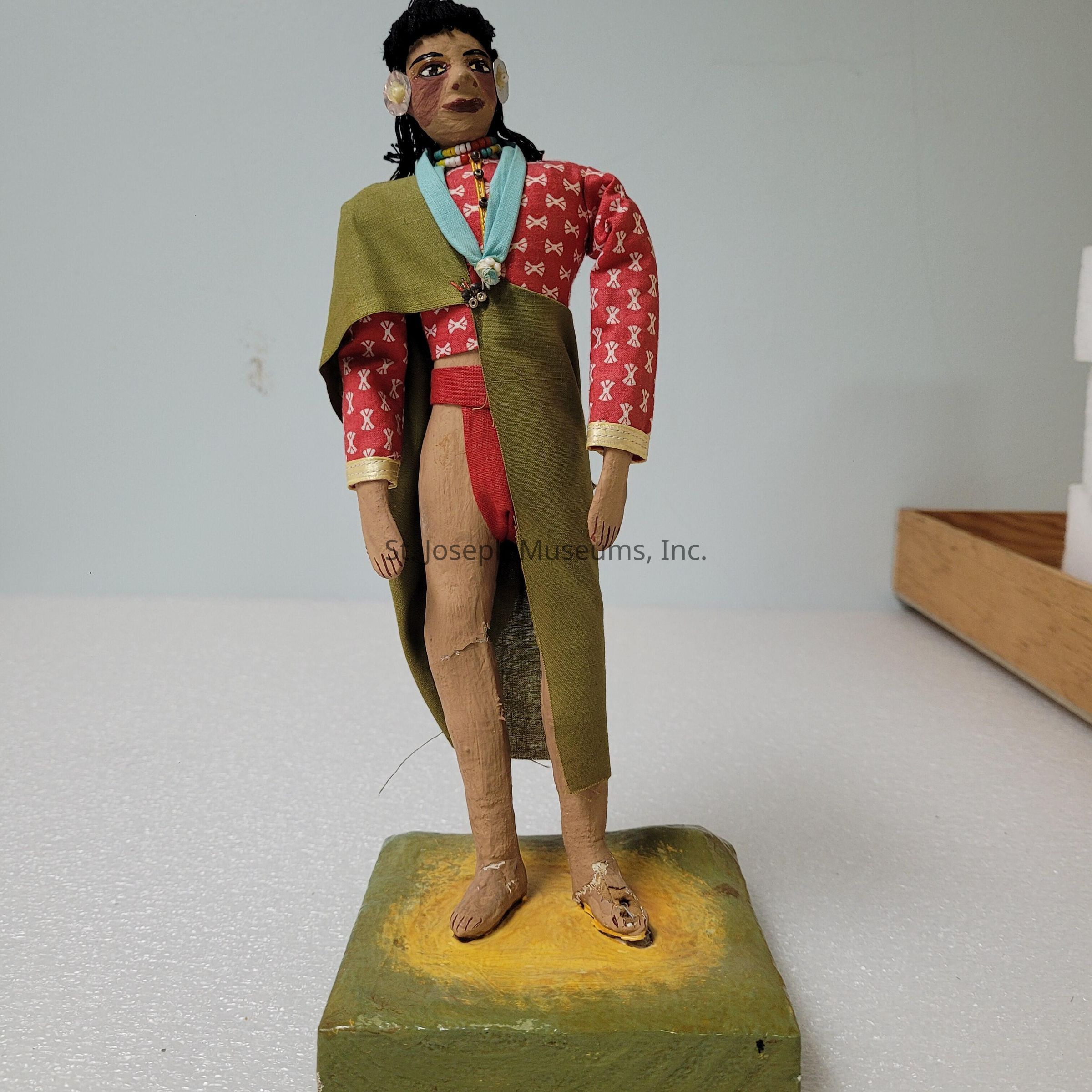 The image depicts a handmade figurine standing on a square, painted base with a textured yellow and green surface. The figure is crafted from wood and adorned with a red patterned shirt featuring white motifs, along with long sleeves. Over the shirt, a green sleeveless cloak is draped across one shoulder, tied with a light blue strip of fabric around the neck. The figure wears a necklace of multi-colored beads, adding a vibrant touch to the ensemble. Its facial features are detailed with painted embellishments, and it has black, textured hair adorned with small decorations on each ear. The skirt-like garment the figure wears is simple, extending below the cloak. The figurine stands barefoot, with intricately carved toes, suggesting an artisanal focus on detail. Its posture is slightly tilted, giving it a lively and dynamic stance. Behind the figurine, a plain background enhances its prominent, colorful presence. The reflection of light suggests a smooth, well-finished surface, highlighting the craftsmanship involved in its creation.