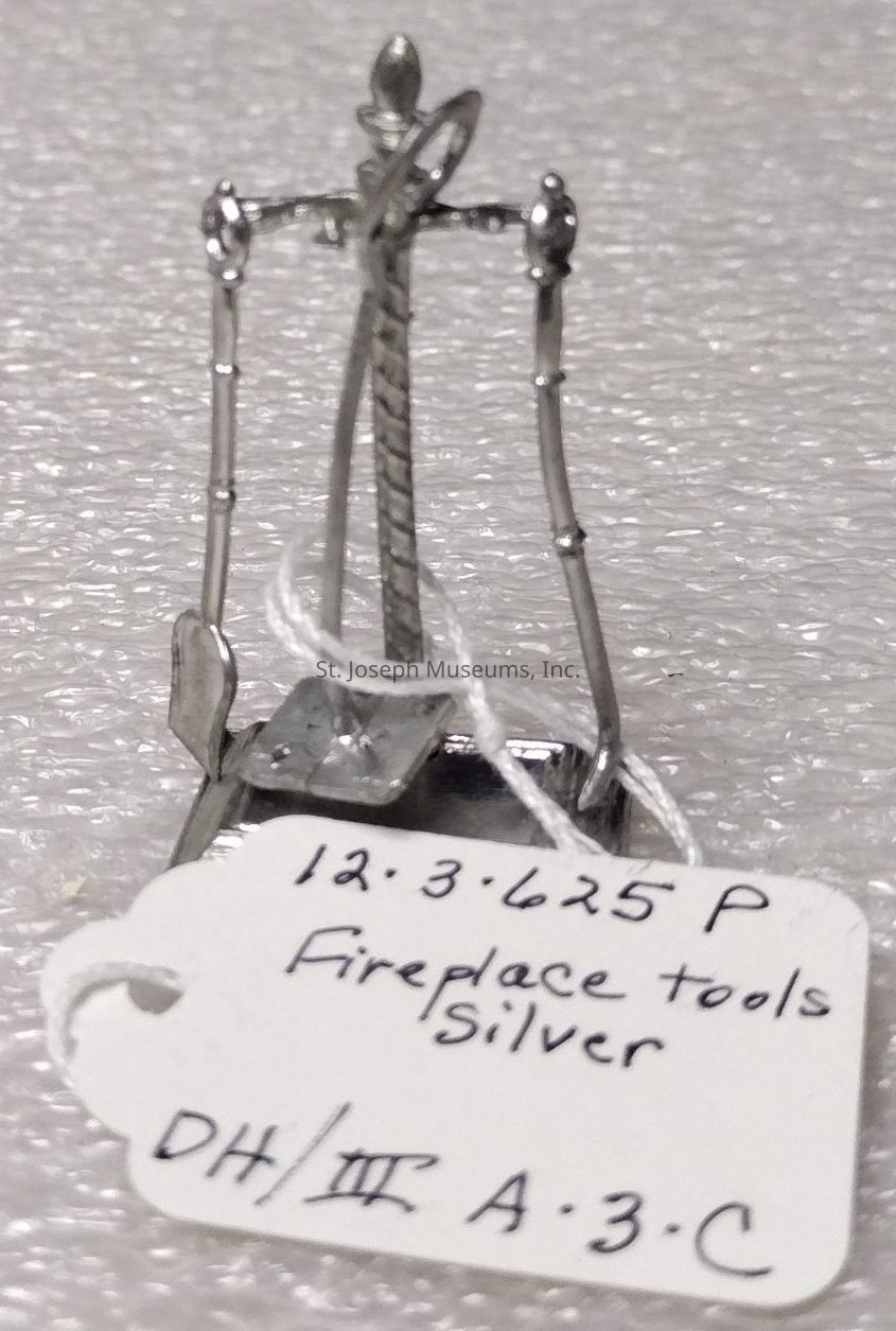 The image shows a miniature set of fireplace tools made from silver, standing on a textured surface. The set includes various tools, each intricately designed with fine detailing that suggests an ornate, decorative style. A handwritten tag is attached to the tool set, inscribed with the text "12.3.625 P, Fireplace tools, Silver, DH/III A.3.C." The tools appear to be delicately crafted, indicating they could be a model or a collectible item. The overall appearance of the set reflects both functionality and artistic craftsmanship, making it a unique piece that blends practical design with aesthetic appeal.