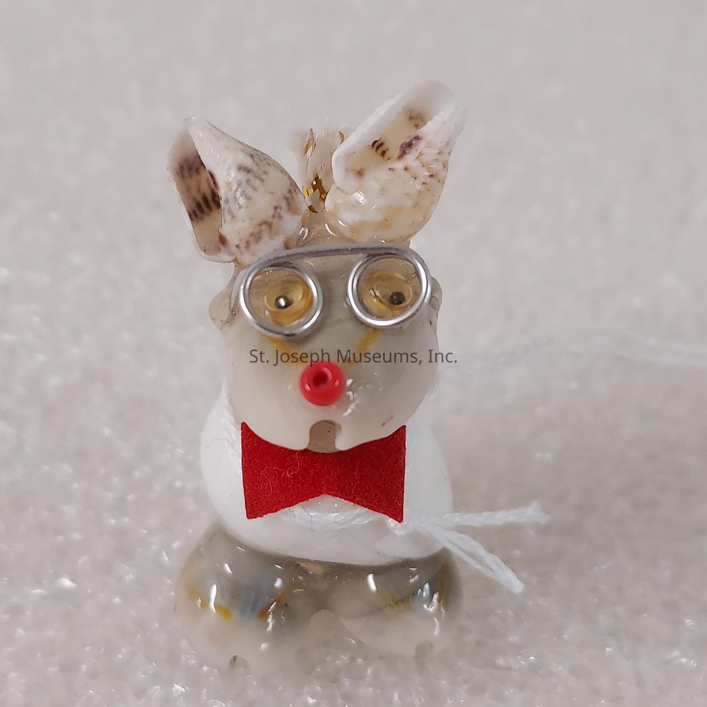 The image depicts a small, whimsical figurine created from seashells, wire, and small decorative elements. The figure resembles a cartoonish rabbit, with seashells forming the ears and body, while coiled wire and beads are used for the eyes. A tiny red bead serves as the nose, enhancing its playful expression, and a miniature red felt bow tie adds a charming touch to its appearance. The entire figure is set against a textured white background, which highlights the intricate details of the handmade sculpture. The figurine conveys a sense of creativity and humor, likely intended as a decorative or novelty item, showcasing resourceful use of natural and simple materials to create an endearing character.