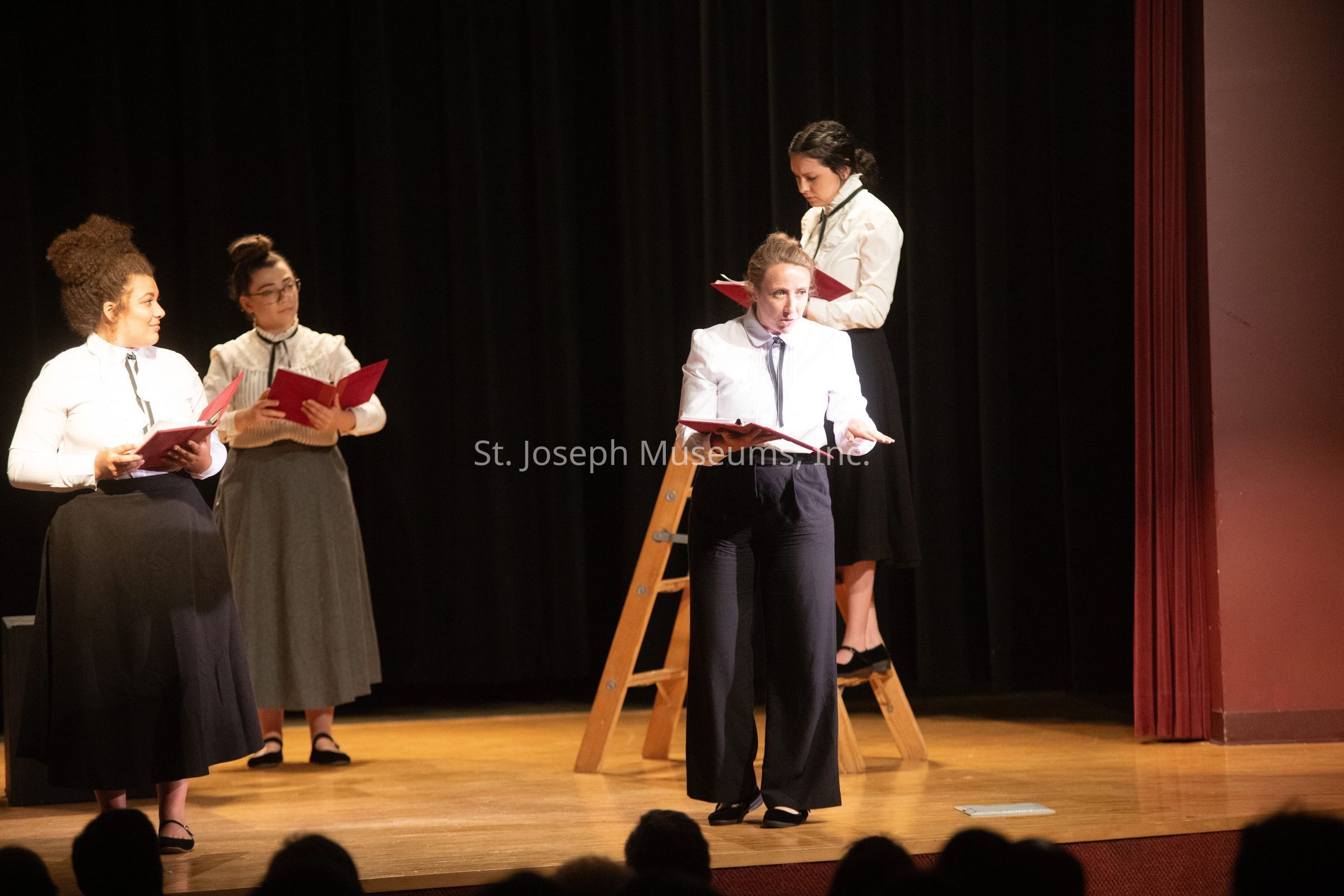 In the image, four individuals are performing on a stage, each holding and engaging with a red script. They are all dressed in formal, period-style attire, with white blouses and dark skirts or trousers. One person stands on a wooden step ladder at the back, focusing intently on her script, while another person at the front gestures expressively as she reads aloud. The dark curtains and spotlights suggest a theatrical setting, creating an intimate and focused atmosphere that highlights the performers' concentration and dedication to their roles, as they deliver their lines to an unseen audience.