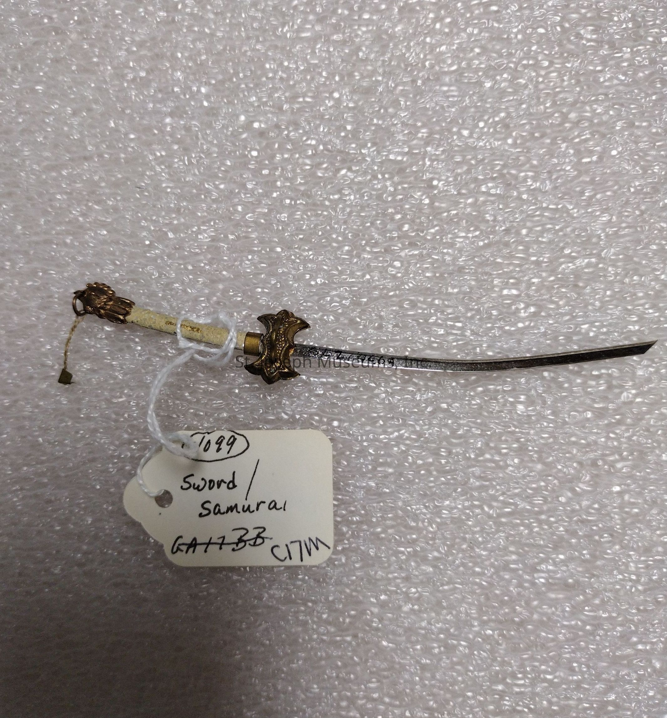 The image features a miniature decorative samurai sword resting on a textured surface, possibly bubble wrap. The sword has intricate detailing on its hilt and guard, with a small tassel hanging from the pommel. A string is attached to the sword, holding a white tag that reads "Sword / Samurai" along with what appears to be catalog numbers or identifiers, such as "1089" and "GA173B." The tag is attached just below the hilt with a string that is looped around the handle. The background consists of evenly spaced bubbles, contributing a subtle texture to the composition, contrasting against the metallic and ornamental details of the sword.
