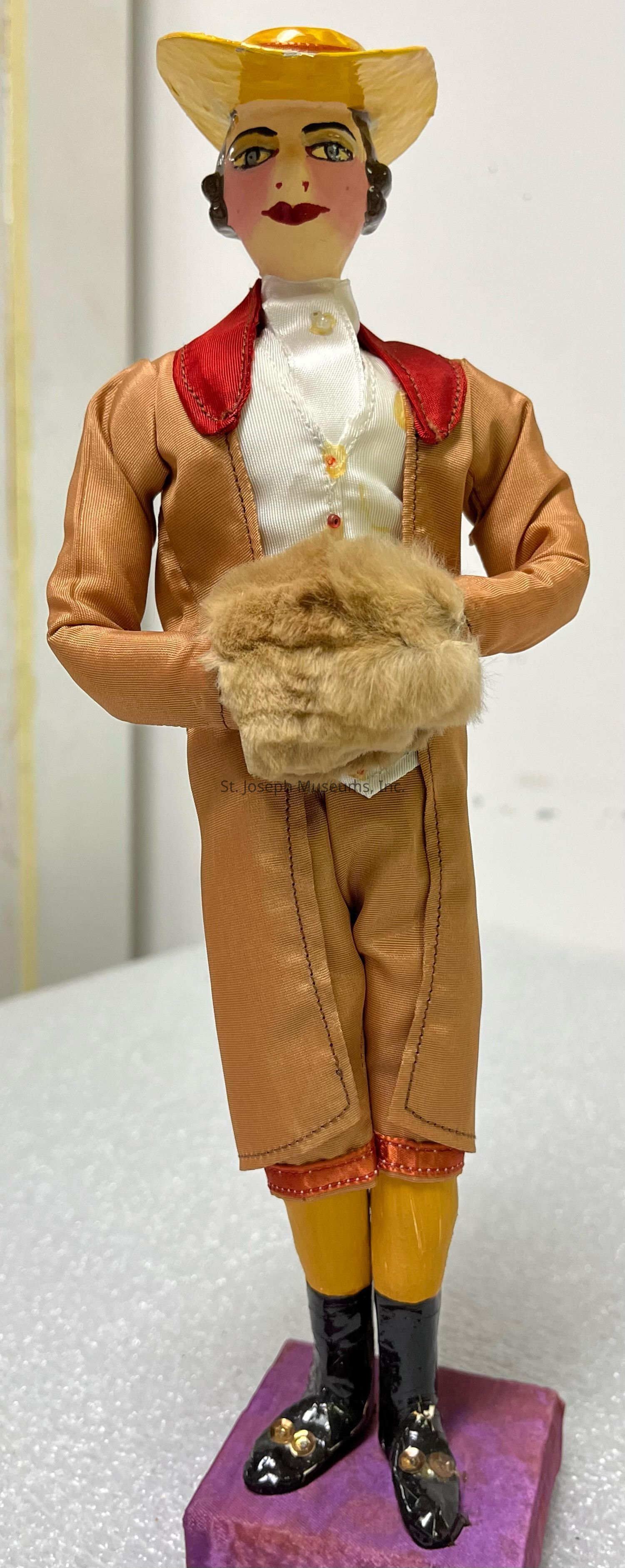 The image depicts a decorative figurine standing atop a purple base, characterized by an intricate outfit reminiscent of formal attire. The figurine wears a brown suit with a white shirt, accented by a red collar and a detailed yellow waistcoat. Its hands hold a fluffy, light brown muff, adding texture to the ensemble. The figure also wears tall, shiny black boots adorned with metallic embellishments, contributing to the overall ornate design. Its face is finely painted, featuring prominent eyebrows, red lips, and vivid eyes, underlined by a sharp gaze. To complete the look, the figurine dons a wide-brimmed yellow hat with a glossy finish. The background of the image is plain, emphasizing the details of the figurine, which exudes an air of elegance and nostalgia, capturing a blend of craftsmanship and artistic expression typically seen in handcrafted collectibles or vintage dolls. This figurine could be a representation of historical or cultural attire, possibly suggesting a character from a specific era or story.