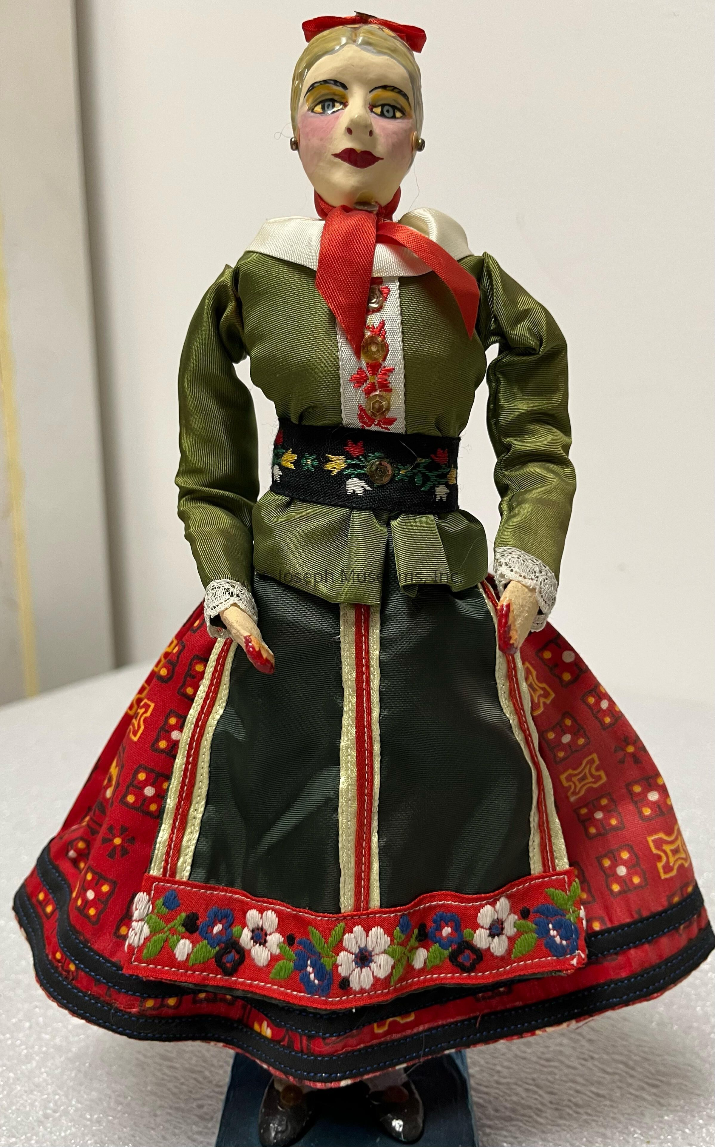 The image depicts a handmade doll dressed in traditional attire, featuring a green blouse with intricate floral embroidery and a prominent white collar accentuated by a red ribbon tied in a bow around the neck. The doll's face is artistically painted, showcasing distinct facial features and a serene expression. Its skirt is richly decorated with vibrant patterns, displaying a combination of red, floral, and geometric designs that add to its cultural significance. The design is further complemented by a black belt with colorful embroidery, adding contrast to the overall appearance. The doll stands poised, with delicate hands, wearing black shoes, and its hair is styled neatly, topped with a small red accessory. Its craftsmanship suggests careful attention to detail, capturing the essence of traditional clothing and artistry. It rests on a textured white surface, highlighting its intricate details and vivid colors against the simple background.