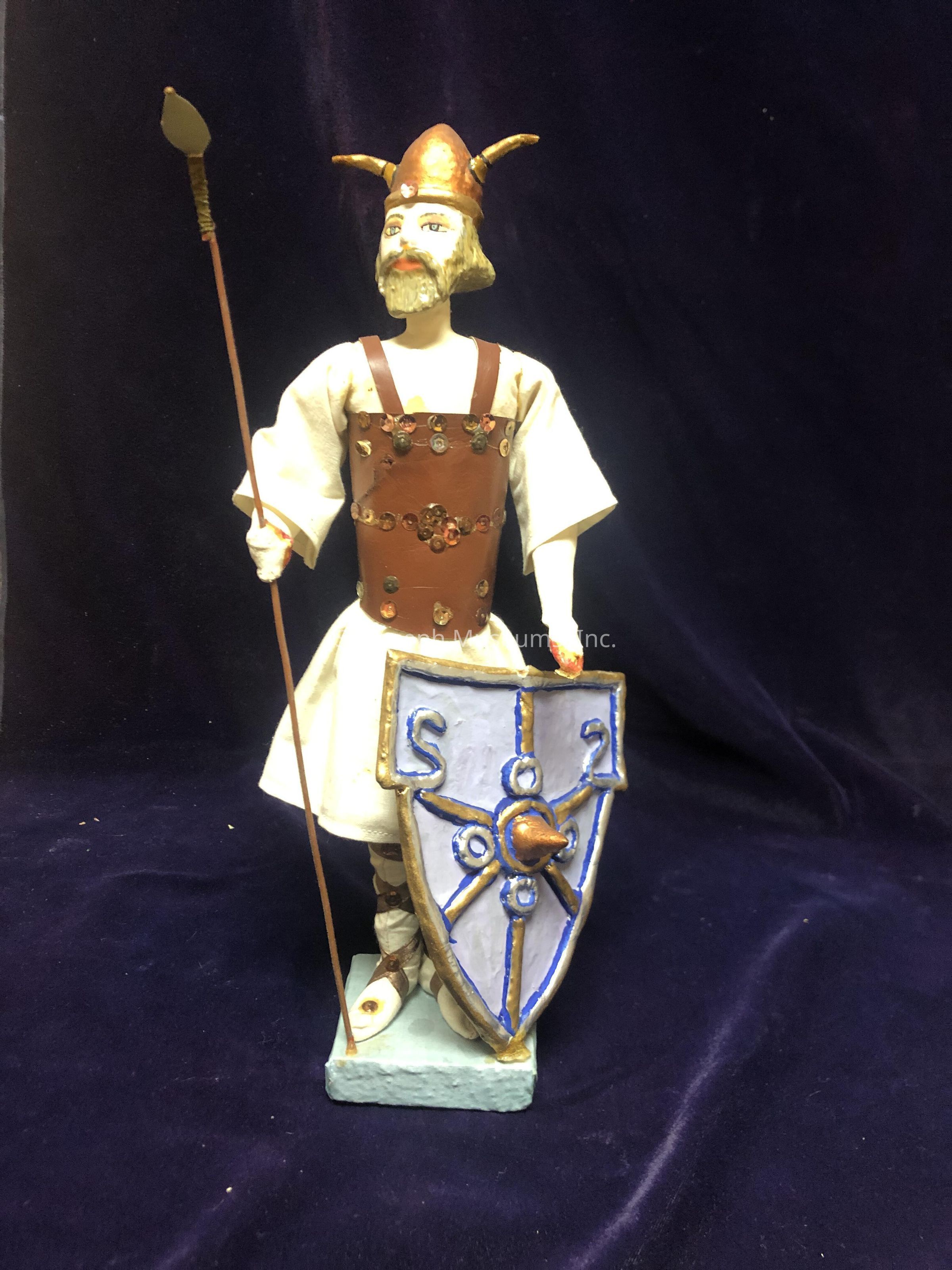 The image shows a figurine of a bearded warrior wearing a helmet adorned with horns and dressed in a white tunic with a brown leather-like vest decorated with ornate buttons. The warrior holds a spear in his right hand and carries an elaborate shield in his left hand, featuring intricate blue and gold designs, perhaps resembling ancient symbols. The figurine stands on a light blue base, set against a dark velvet backdrop, giving the scene an impression of a historical or mythical representation, evoking thoughts of bravery and valor from a bygone era, and capturing attention with its detailed craftsmanship and imaginative presentation.