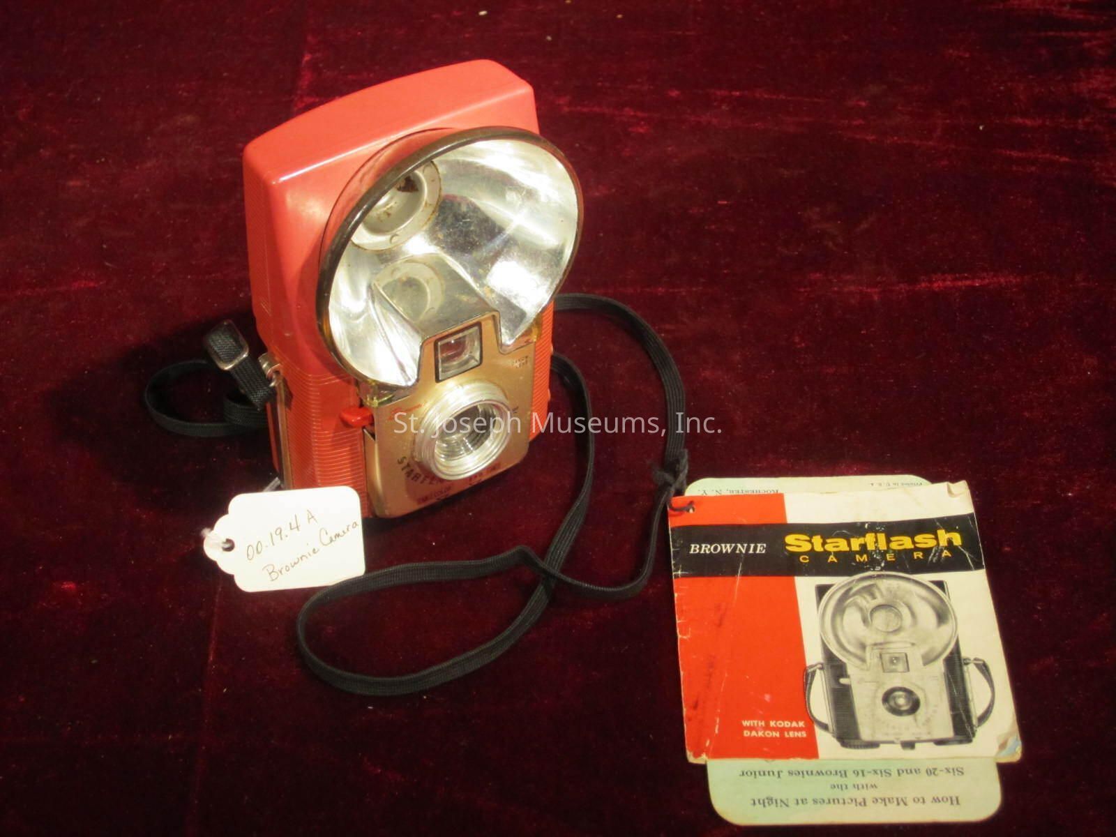 The image depicts a vintage camera set on a dark red surface. The camera has a bright red and metallic gold body, with a prominent flashbulb component at the top. A handwritten tag attached to the camera reads "00-19.4 A Brownie Camera." To the right of the camera lies a user manual or instruction booklet with a cover featuring an illustration of the same camera model and "Brownie Starlash Camera" written on it. The booklet is slightly worn, suggesting age and use. The overall scene evokes a sense of nostalgia, capturing the appeal of classic photographic equipment.
