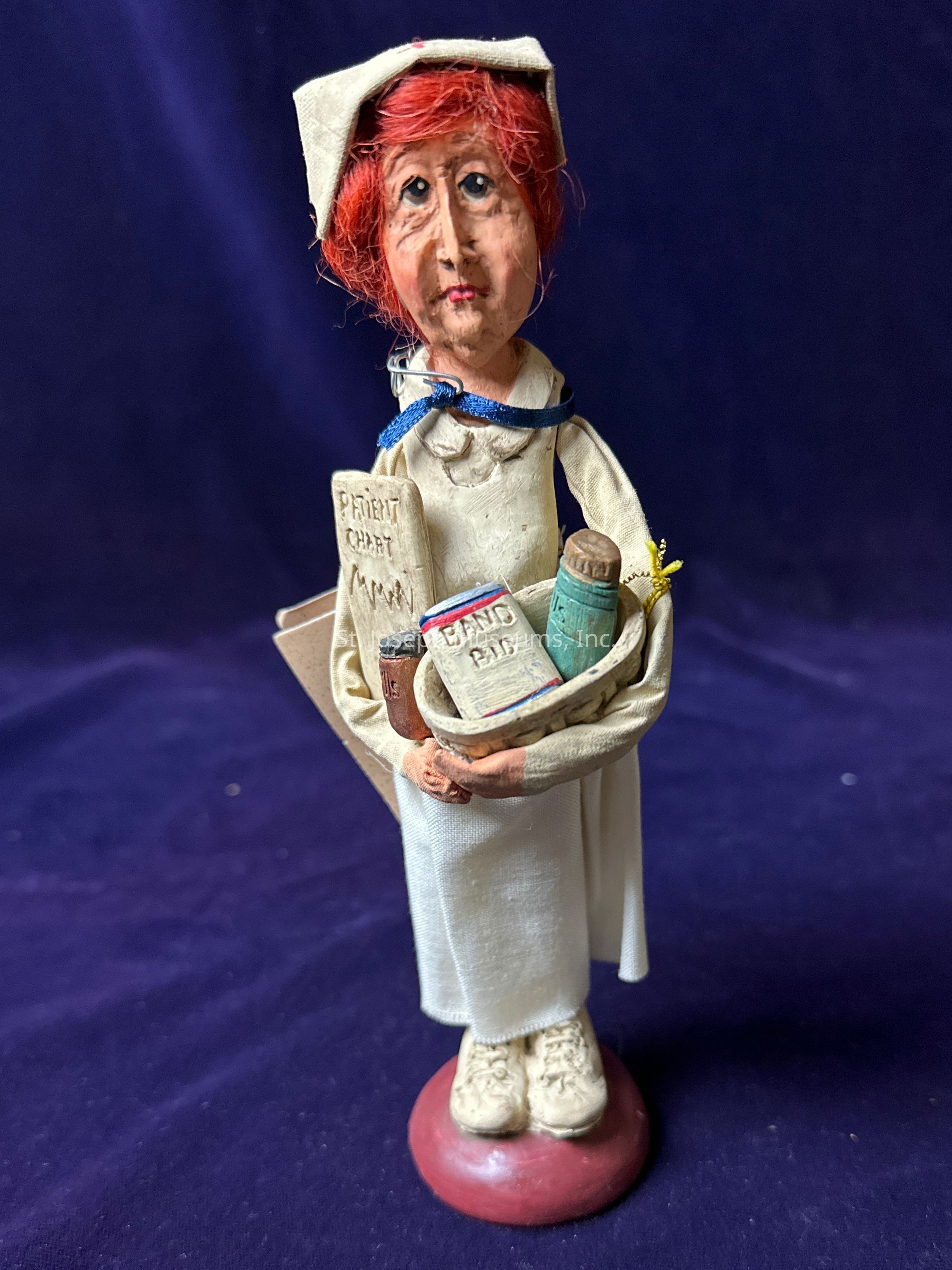 The image shows a figurine of a nurse with a whimsical, handcrafted appearance, featuring distinctive facial expressions and red hair under a traditional nurse's cap. The nurse is adorned in a white uniform, complete with a blue ribbon and shoes, standing on a red base against a dark background. She holds a tiny detailed armful of items including a patient chart, a book labeled 'Band Aid', and additional objects that resemble medical supplies or scrolls. The intricate design and the hand-painted features contribute to the unique charm and character of the figure, evoking a nostalgic and artistic representation of the nursing profession.
