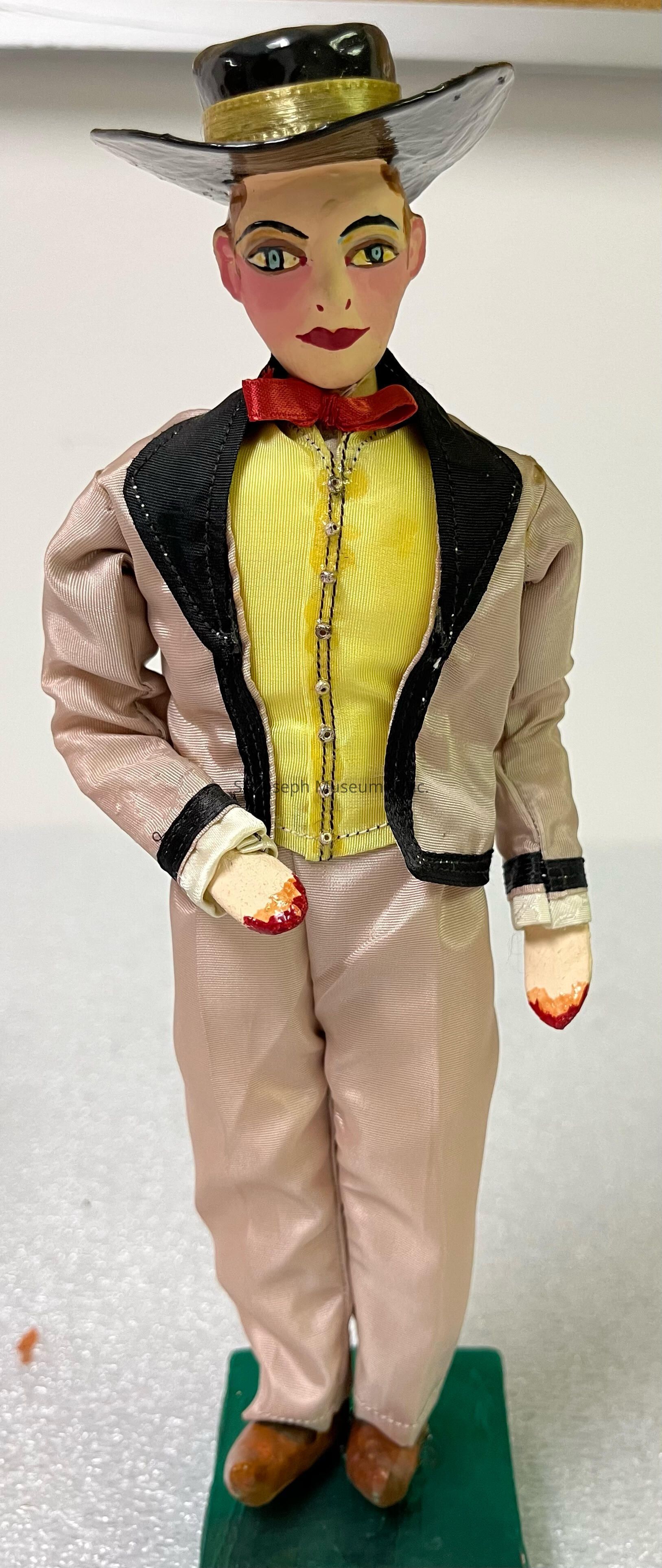 The image shows a small figurine of a person dressed in an outfit reminiscent of formal or traditional attire, featuring a yellow shirt under a black-bordered jacket, beige pants, and a wide-brimmed hat with a dark bend crowned by a band. The figure’s face has exaggerated and stylized features, including large painted eyes and red lips, while the hands appear to be painted with a red detail at the tips, possibly representing gloves or a stylistic choice. The figure is mounted on a green rectangular base, standing with an air of poise.