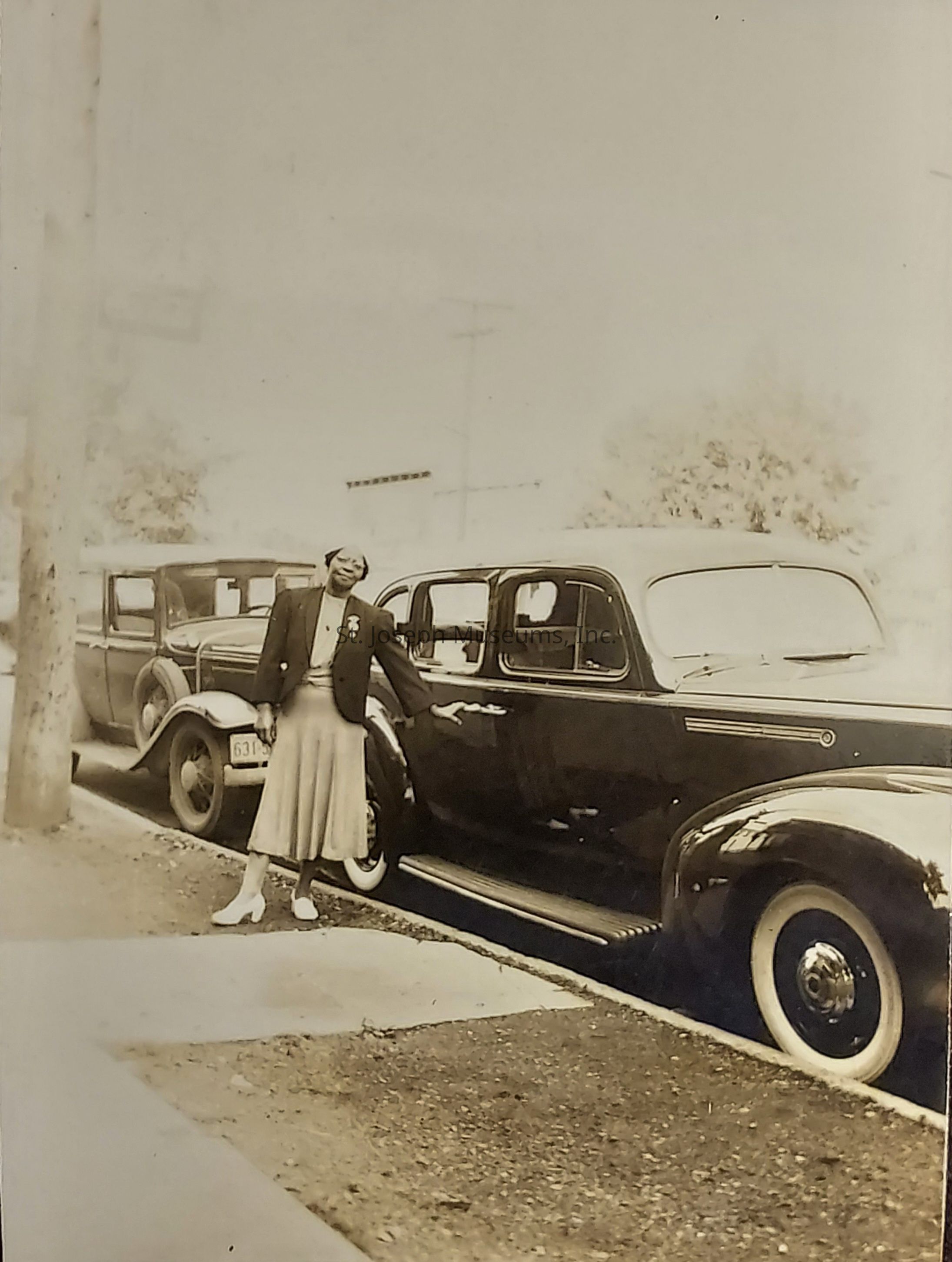In this vintage photograph, a woman stands next to a sleek, classic car parked along the street, dressed in mid-20th-century attire with a neatly tailored jacket and a calf-length skirt. She strikes a proud pose beside the shiny vehicle, which boasts elegant lines and distinct white-wall tires. The background features another car parked behind, with blurred trees and telephone poles indicating a suburban or urban setting of that era. The scene is composed in sepia tones, adding an air of nostalgia and giving a glimpse into a moment of everyday life from a bygone time, capturing the essence and style of a past decade.