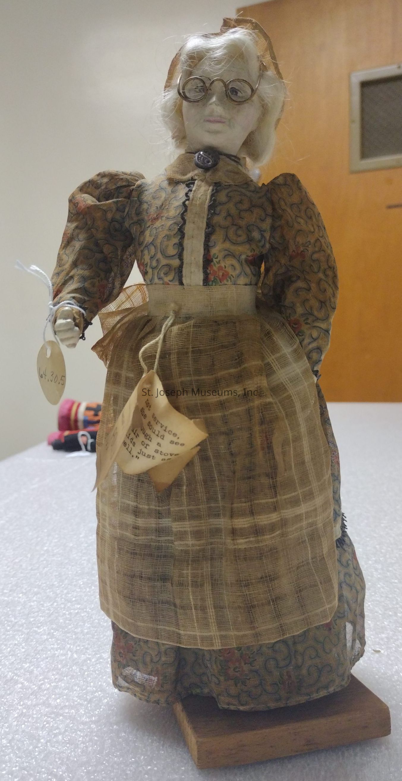 The image depicts a vintage doll standing upright on a wooden base, adorned in an old-fashioned outfit consisting of a patterned dress with puffed sleeves and a plaid apron tied around its waist. The doll has a pale face with wire-rimmed glasses perched on its nose and wears a small, decorative button at the collar. Its white hair is styled in waves, adding to the antique look. Attached to the doll's hand and apron are tags, one of which appears to contain typewritten text, further enhancing its historical appearance. In the background, there is a wooden door with a metal mesh panel, suggesting the setting might be a room or display area.