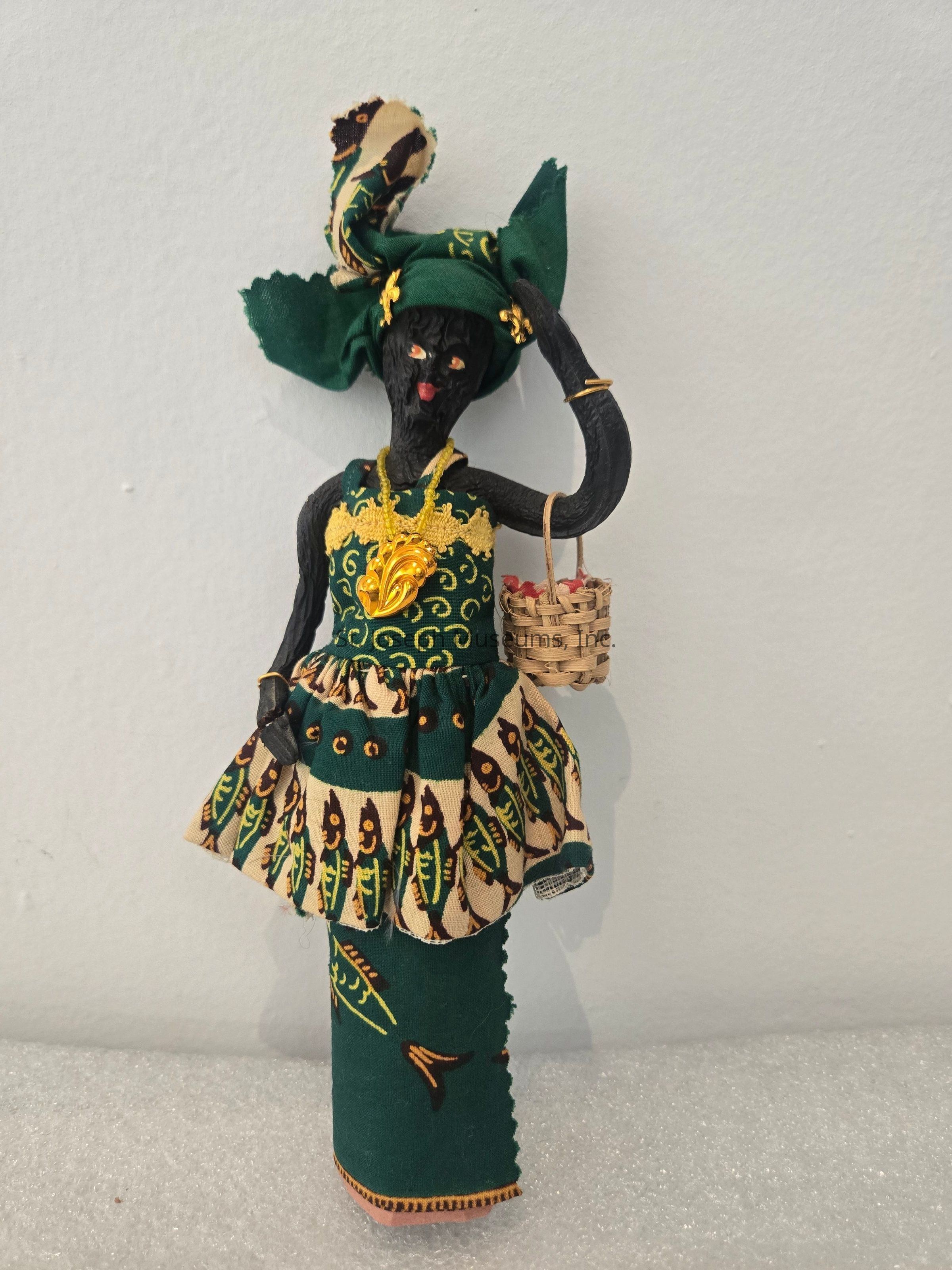 The image shows a handcrafted figurine wearing a vibrant, green, and patterned outfit adorned with a headwrap that completes its traditional attire, displaying intricate fish designs in its clothing. The figure holds a small basket filled with tiny red items, adding an element of purpose or task. The combination of textures and patterns on the clothing stands out against the plain background, focusing attention on the detailed craftsmanship. The bright gold necklace and matching accent on the dress further embellish the figure, suggesting cultural or festive significance. The posture suggests a poised, graceful stance, giving the figure an air of dignity and elegance.