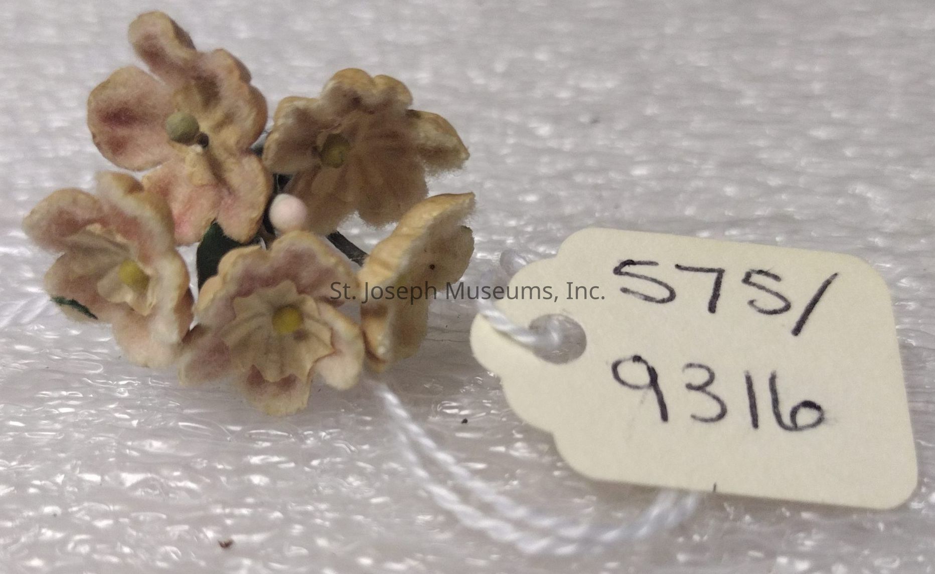 The image displays a small cluster of delicate, light pink artificial flowers with subtle yellow centers, resting on a textured, bubble wrap surface. Attached to the floral arrangement is a white string holding a cream-colored tag, which bears the numbers "575/9316" written in black ink. The overall composition portrays a sense of simplicity and quaintness, suggesting that the flowers might be part of a decorative piece or a keepsake. The background's bubbled texture contrasts with the smoothness of the tag and the soft appearance of the flowers, adding depth to the visual presentation.