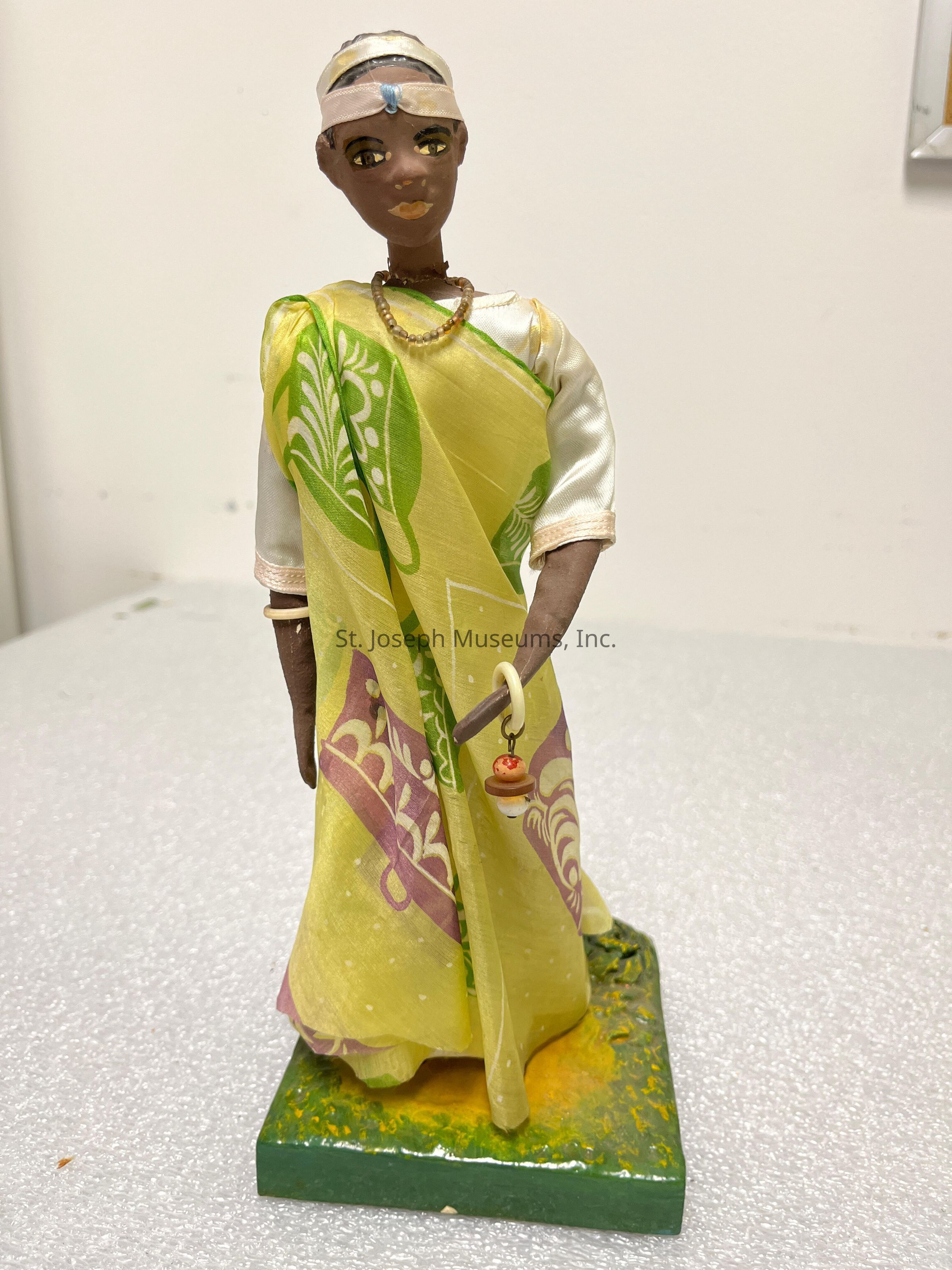 The image displays a handmade figurine of a person adorned in traditional attire. The figure wears a colorful, wrapped garment with an intricate pattern in shades of green, yellow, and purple. The facial expression is detailed, featuring large, expressive eyes and a subtle smile. A small headpiece and necklace add to the authenticity of the traditional attire. The figure is positioned on a rectangular base with a textured surface colored in green and yellow, providing stability and an element of artistry. The right hand of the figure holds a small ornament that appears to be a symbolic accessory. This carefully crafted figure stands against a neutral background, emphasizing its vivid colors and the cultural significance of its attire.
