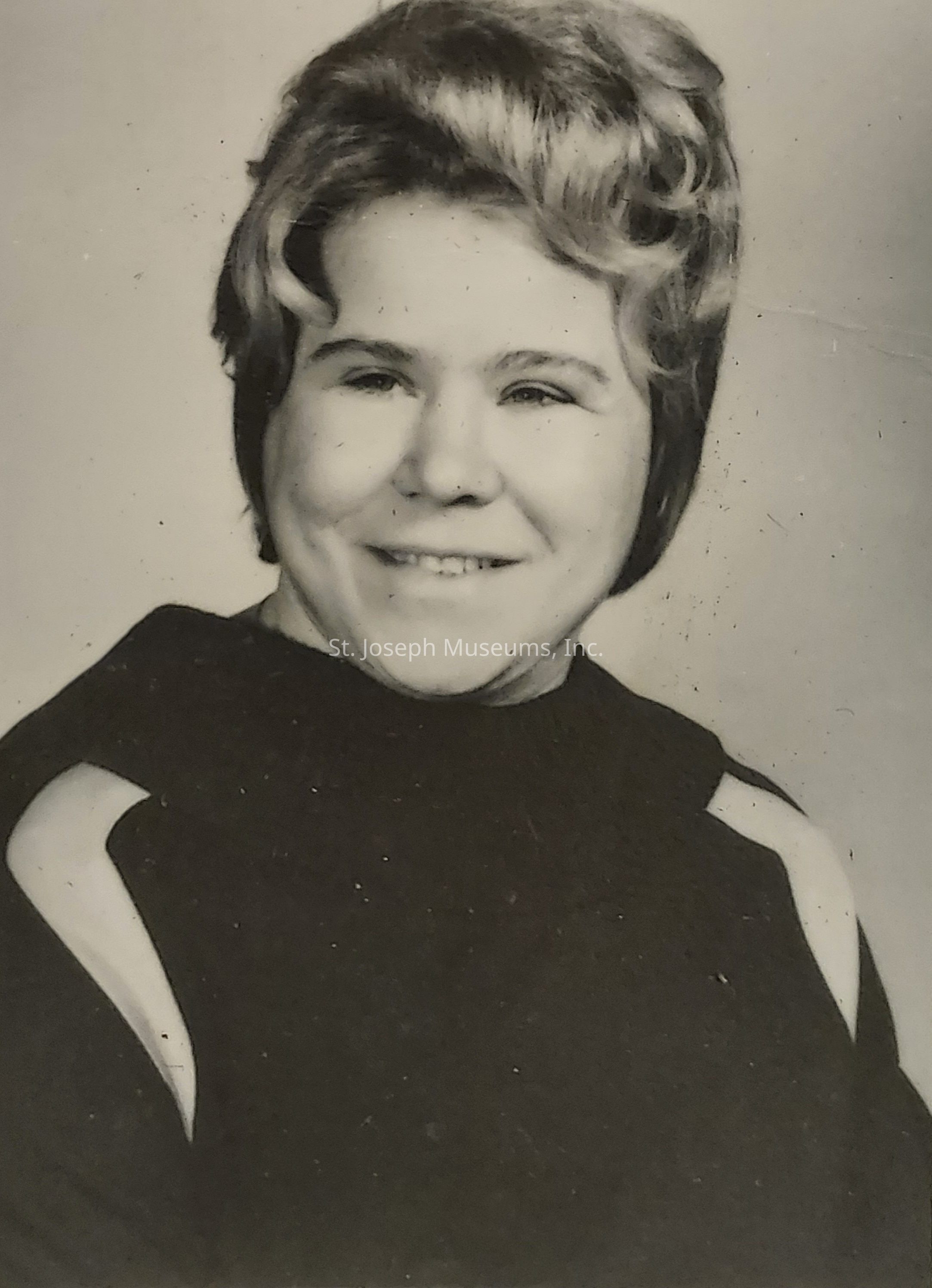 The image depicts a black-and-white portrait of a person who is smiling gently, dressed in a dark top with distinctive white stripes on the shoulders, and has hair that is styled neatly, with soft waves adding a touch of elegance to the overall appearance; the photograph appears to be aged, conveying a sense of nostalgia, and is characterized by a few visible specks, likely due to time, which contribute to the vintage look, further highlighting the timeless nature of the portrait as the person's serene expression and well-arranged hairstyle resonate with a charming and classic aura inherent in photos from the past, evoking a feeling of warmth and historical significance, as viewers might imagine the stories and moments captured during that era, revealing insights into personal histories and the fashion of the time period, making it a fascinating glimpse into the life and times captured within this singular, enduring image.