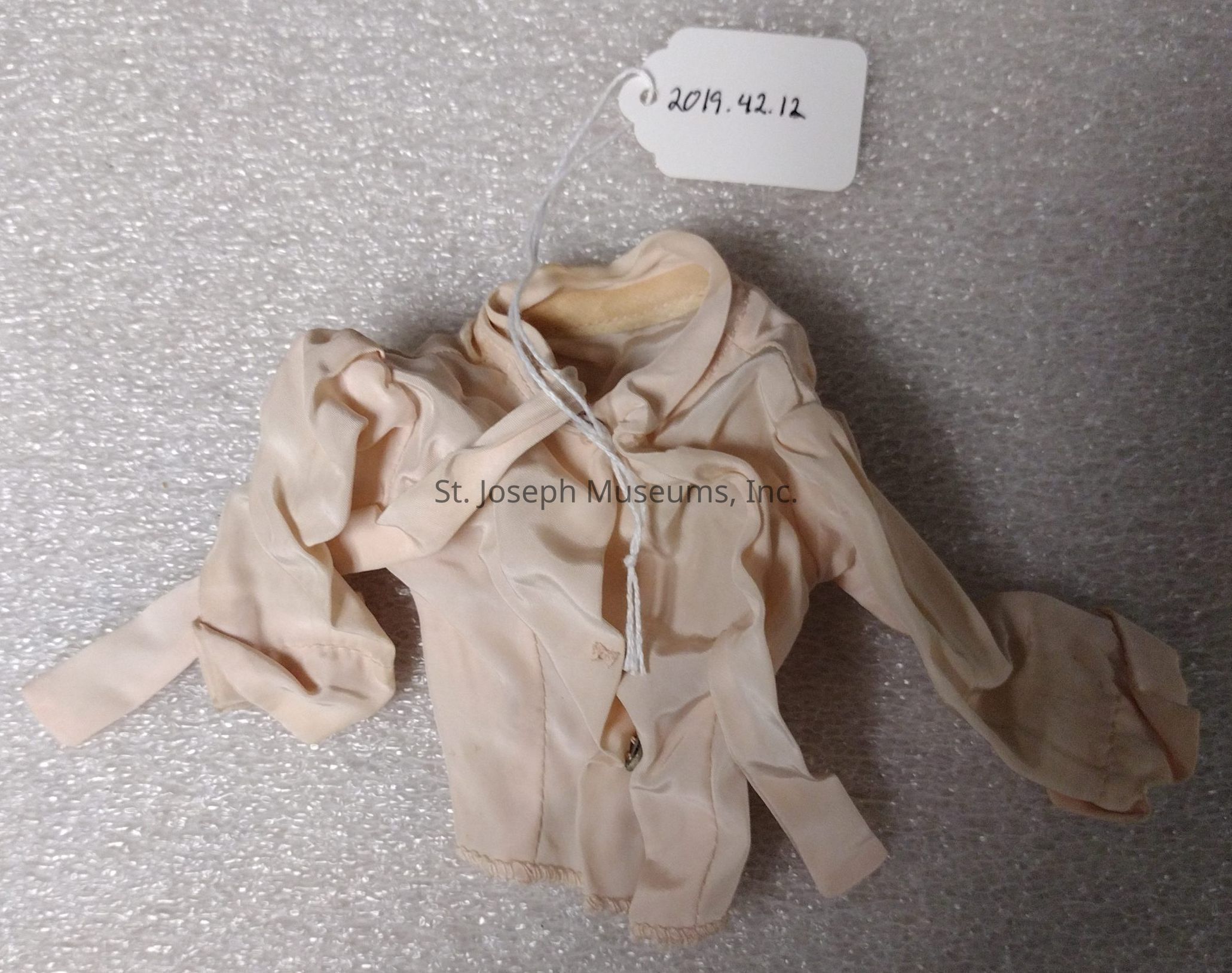 The image depicts a small, cream-colored doll's jacket with long sleeves and a slightly ruffled design, displayed on a textured, foam-like background. The jacket has a high collar and is adorned with a delicate button detail at the front. A white tag is attached to the garment with a string, bearing the inscription "2019.42.12" in black ink, suggesting it might be part of a collection or inventory. The folds and creases of the fabric are visible, indicating a soft, pliable material. The overall appearance of the jacket suggests it is vintage or historically styled, likely intended for a doll or miniature display.