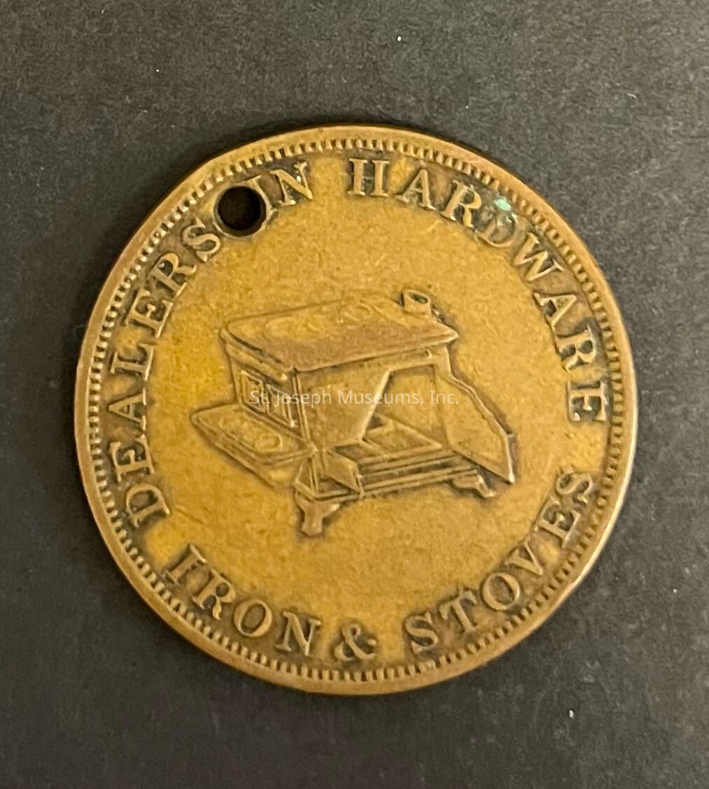 The image displays a round, antique token or medal with a hole near the edge, suggesting it might have been used as a trade token or promotional item. It features an embossed image of a stove in the center, indicative of its association with heating or cooking appliances. Surrounding the stove image are the words "DEALERS IN HARDWARE" and "IRON & STOVES," inscribed in a slightly raised, capitalized font along the border. The token has a brass or bronze appearance, contributing to its vintage look, with subtle wear marks and tarnish.