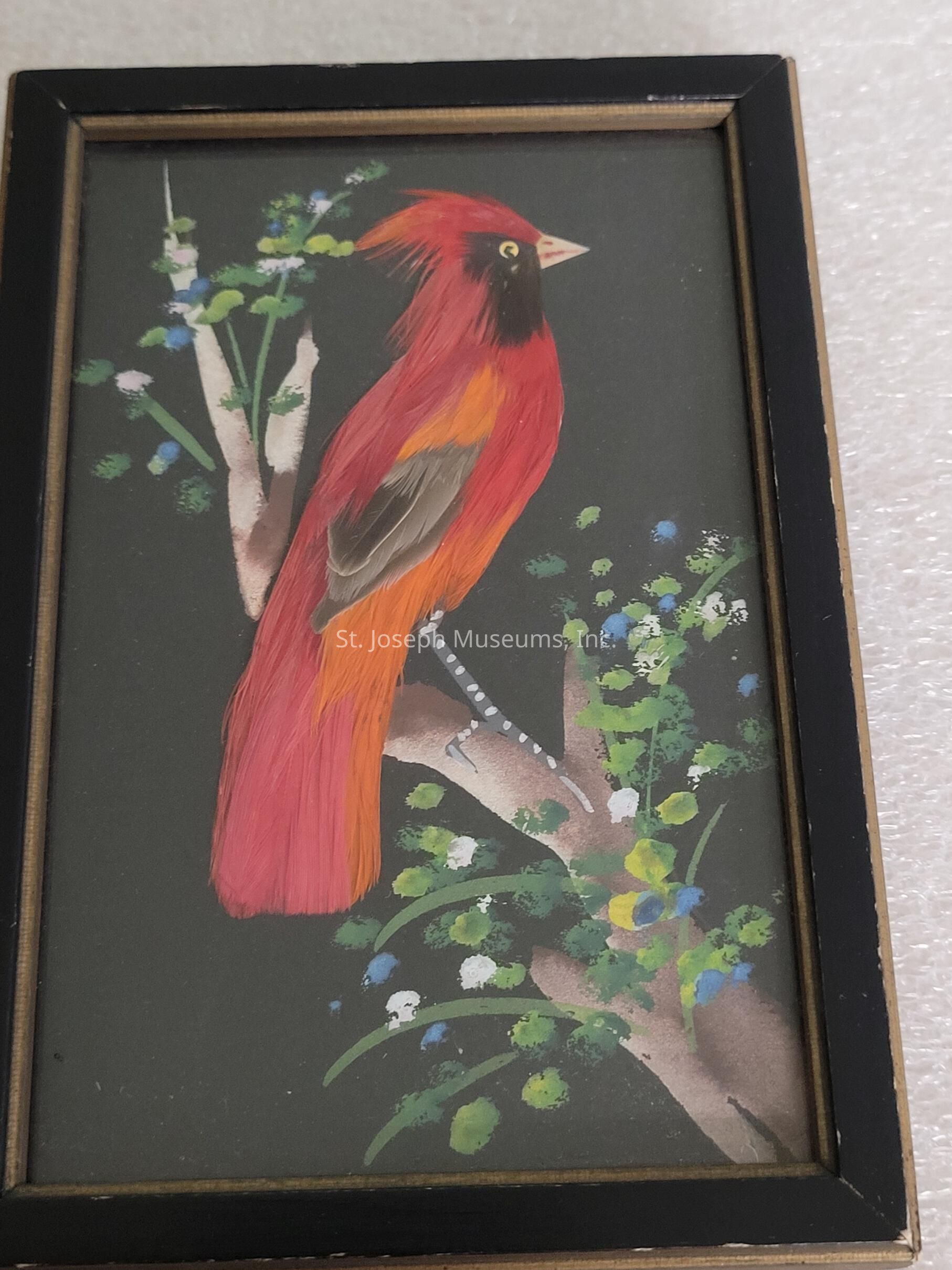 The image showcases a framed artwork depicting a vibrant bird perched on a branch, set against a dark background. The bird is rendered in striking shades of red and orange, with detailed feathers that enhance its lively appearance. Its eye is sharp and observant, while its beak is a pale color, contrasting with the darker features around the face. The branch is adorned with leaves and flowers in hues of green, blue, and yellow, adding an element of natural beauty to the composition. The artwork is enclosed within a thin black frame, lending a sense of completeness to the overall presentation.
